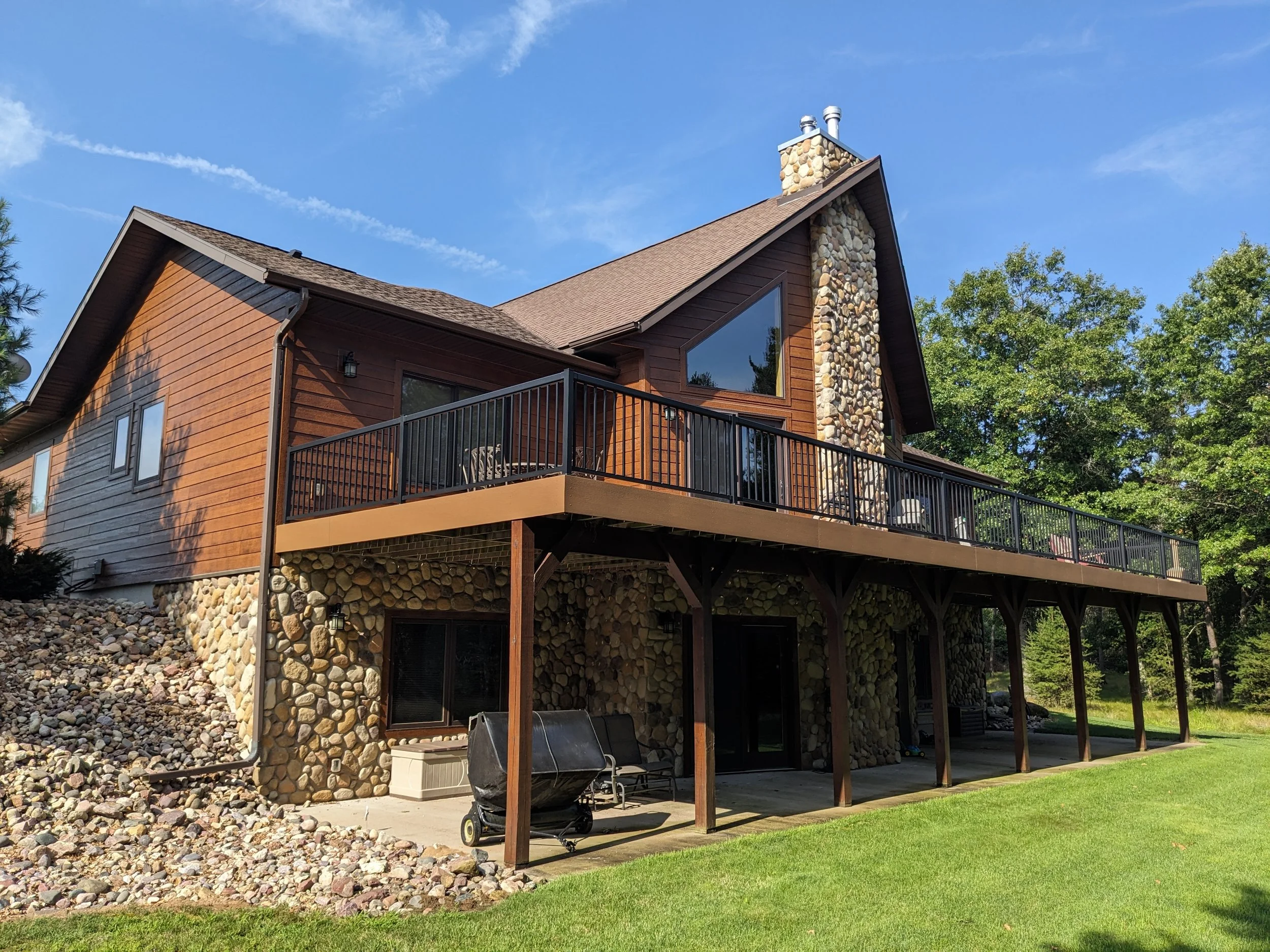 A house with a wooden exterior, stone accents, and a large raised deck with outdoor furniture, surrounded by green grass and trees, under a blue sky.
