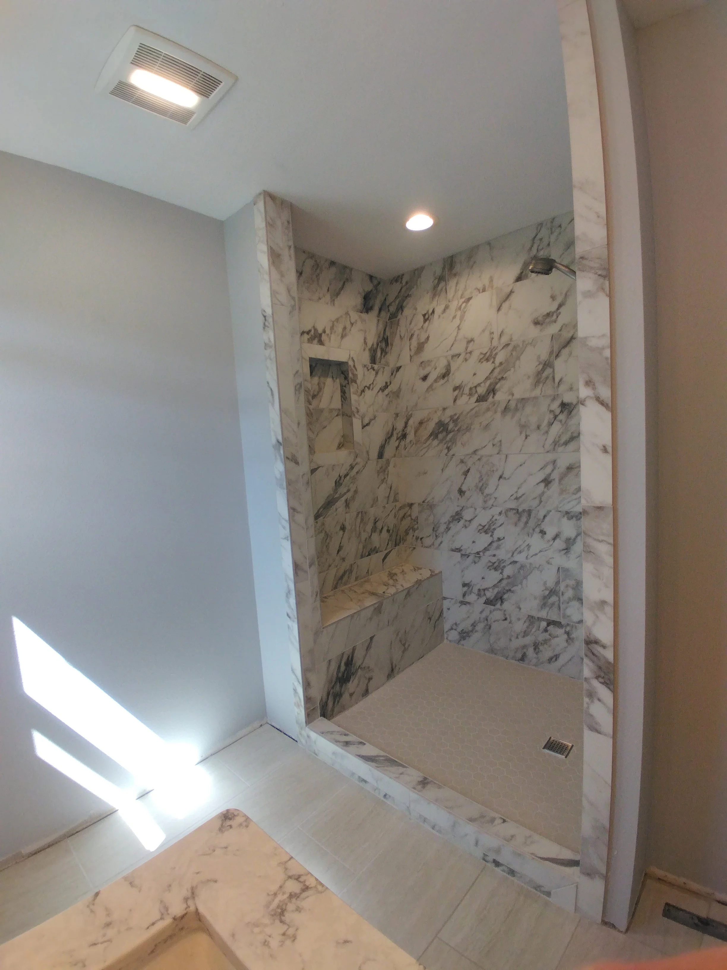 Bathroom shower area with marble tiles, built-in bench, and a small window. The shower has a handheld showerhead and a drain on the floor.