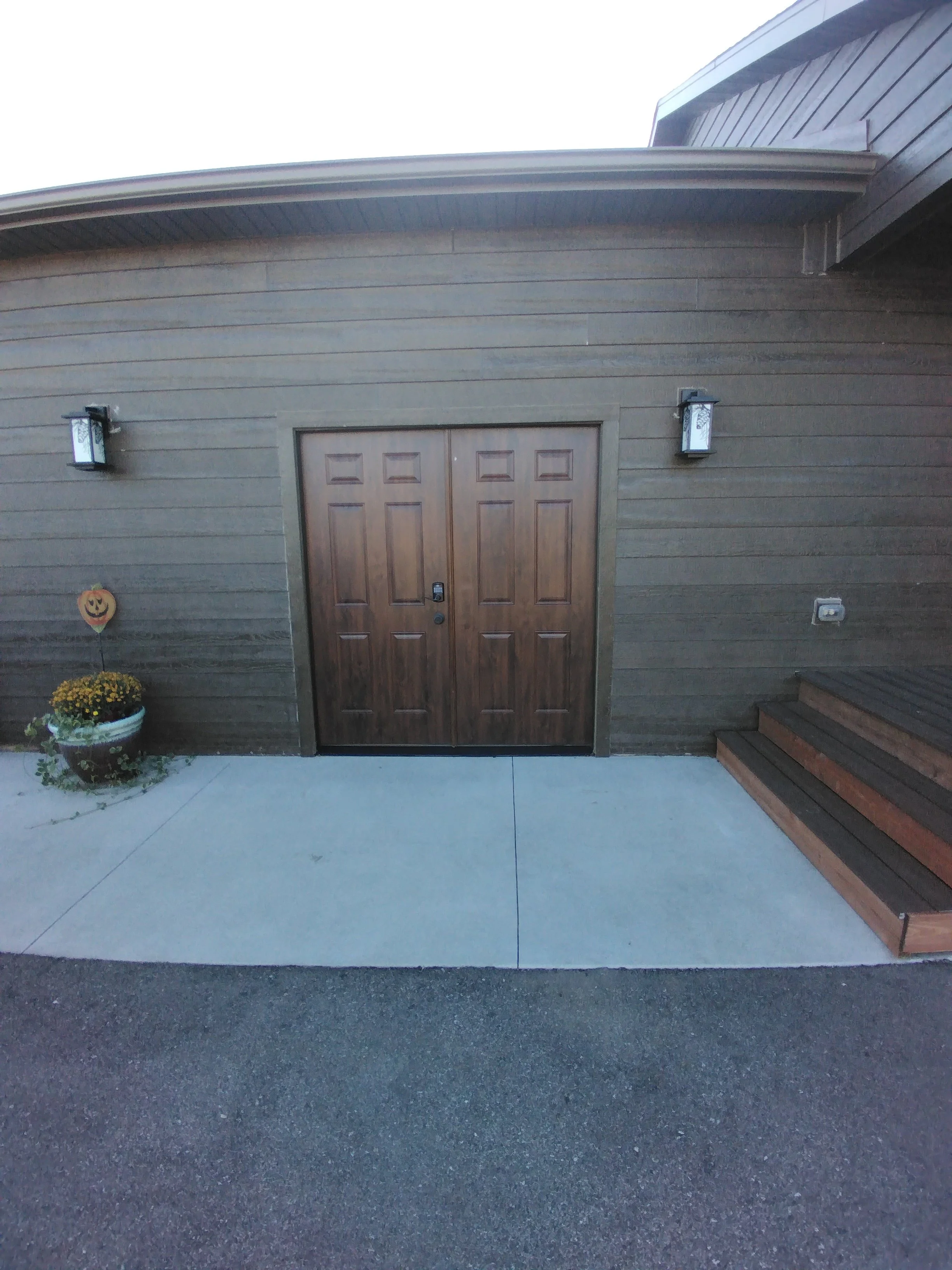 Front view of a house entrance with double wooden doors, outdoor wall-mounted lanterns, a Halloween decoration in a flower pot, and a small wooden staircase.