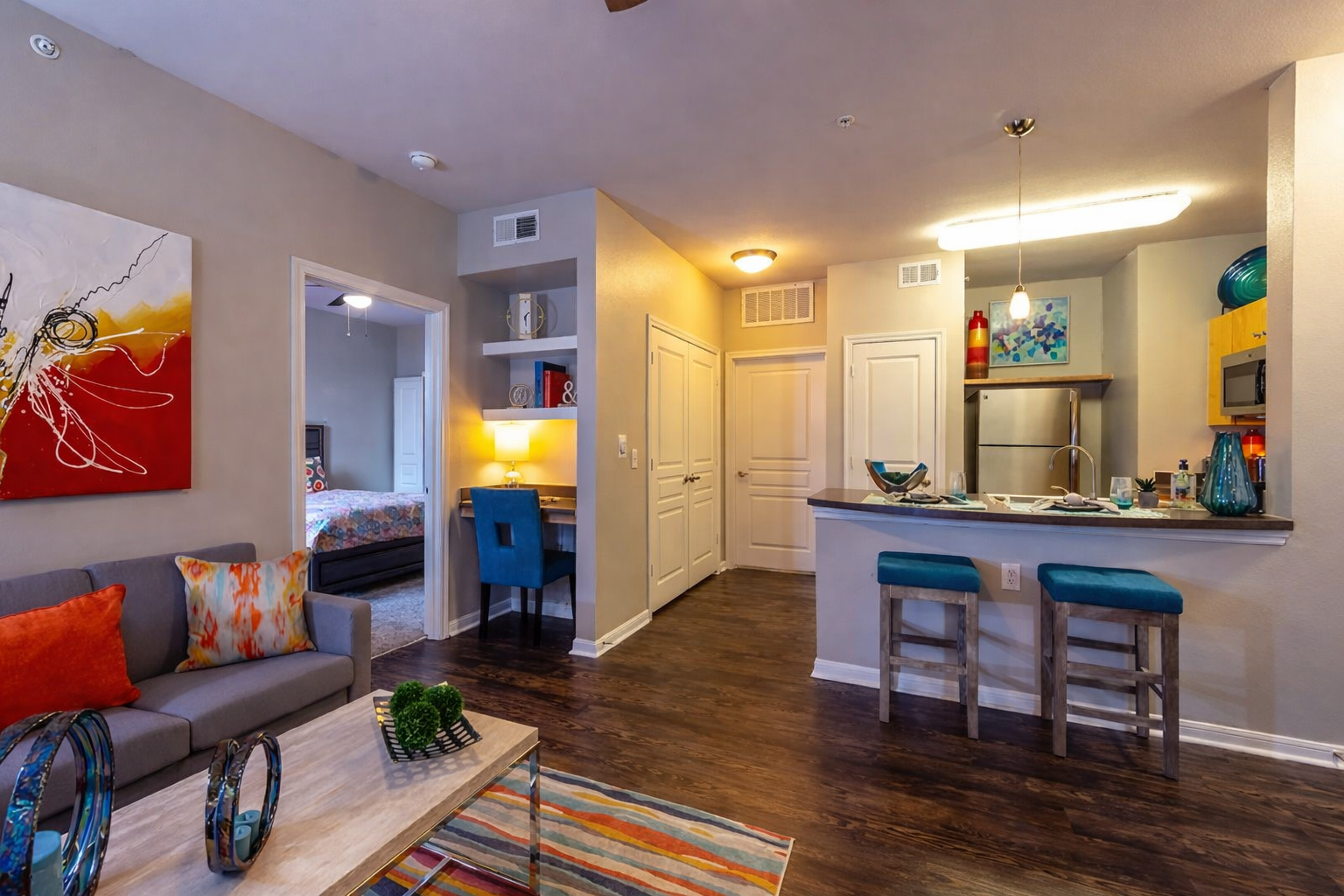 A living room with a gray sofa with orange throw pillows, a colorful abstract painting on the wall, a small table with green plants, a multicolor striped rug, an open kitchen with a breakfast bar and turquoise chairs, and a hallway leading to a bedro