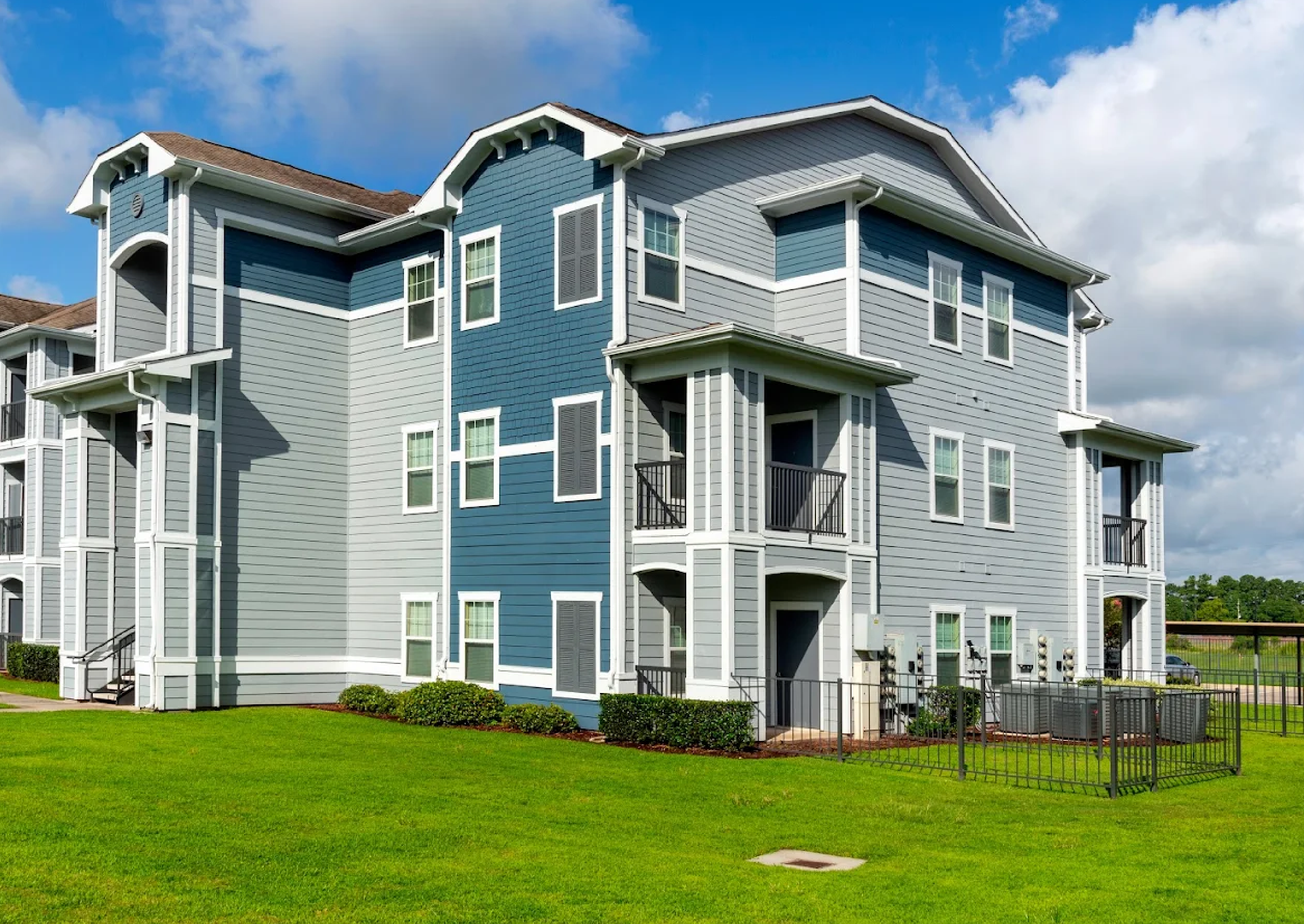 Three-story apartment building with blue and gray siding, white trim, and small balconies, set against a partly cloudy sky, with a well-maintained lawn and a black fence around utility area.