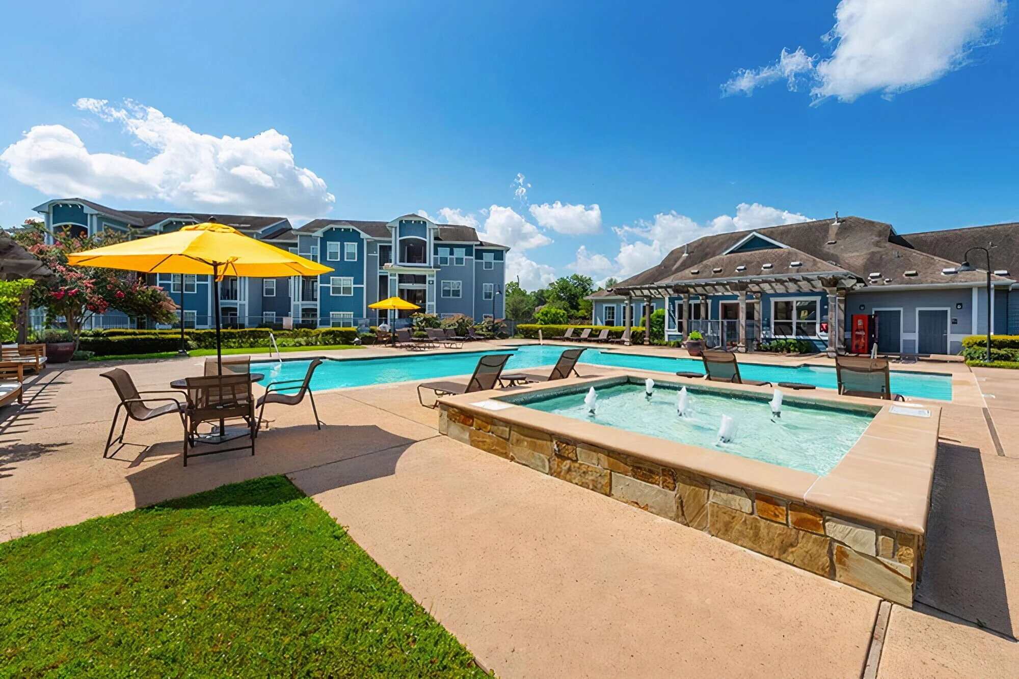 Outdoor swimming pool area with lounge chairs, umbrellas, and a hot tub, surrounded by apartment buildings and a clear blue sky.