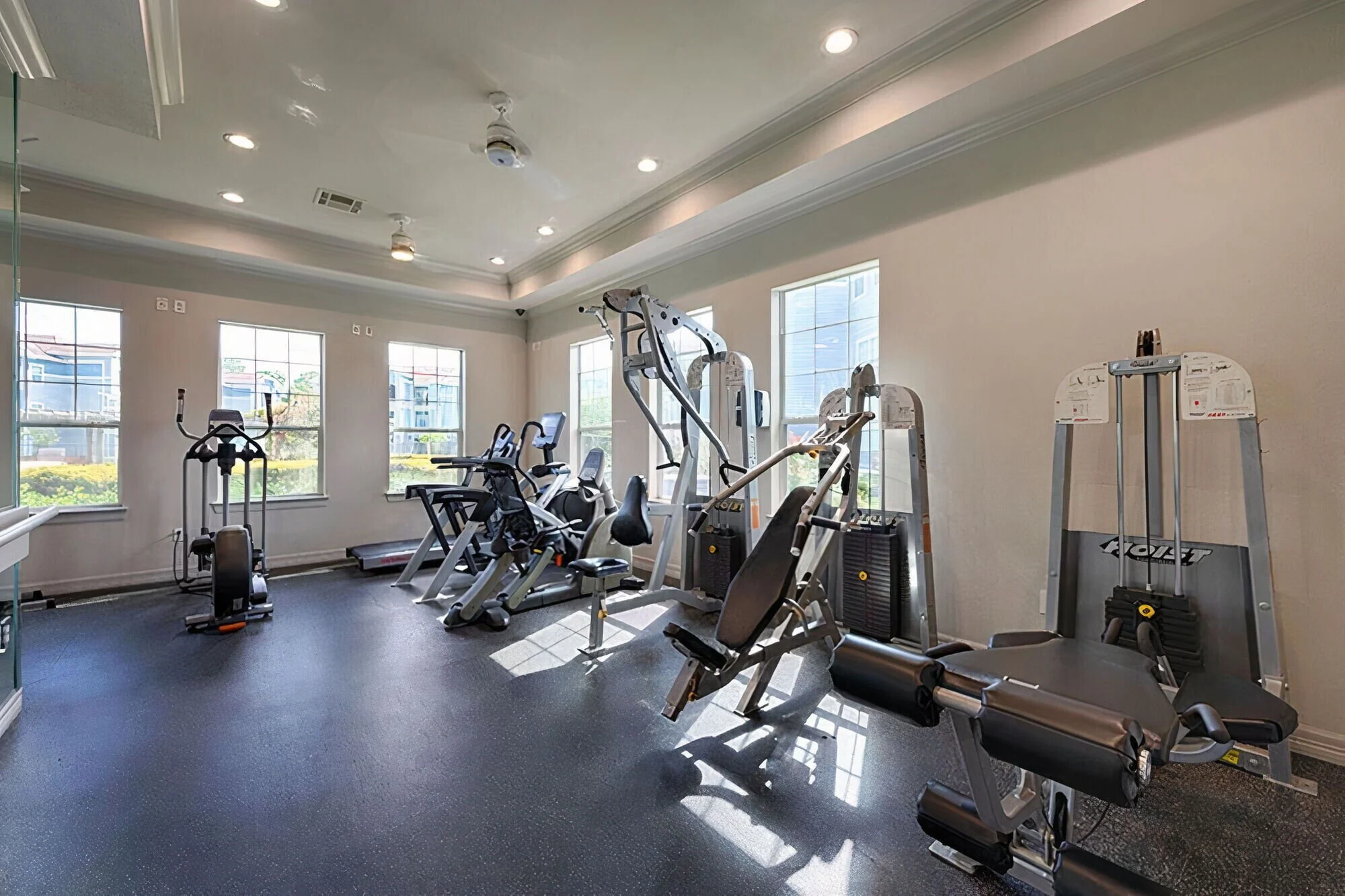 Empty small gym room with exercise machines including a treadmill, elliptical, and weight machines, illuminated by natural light through large windows.