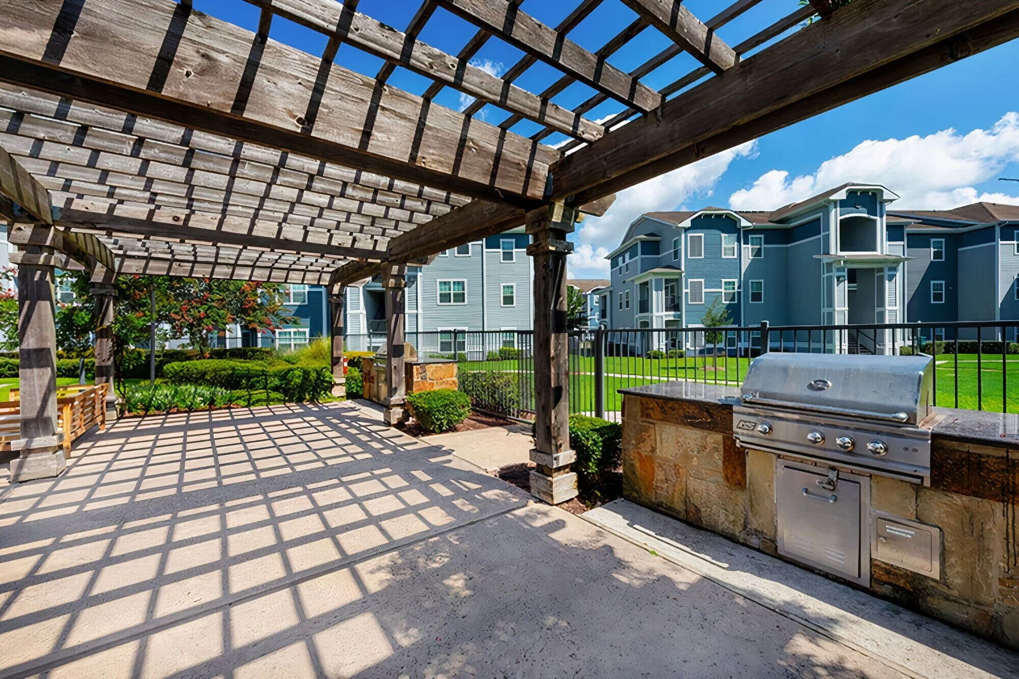 An outdoor communal area in a residential apartment complex with a wooden pergola, concrete flooring, a stainless steel barbecue grill, benches, green grass, and modern multi-story blue apartment buildings in the background under a partly cloudy sky.