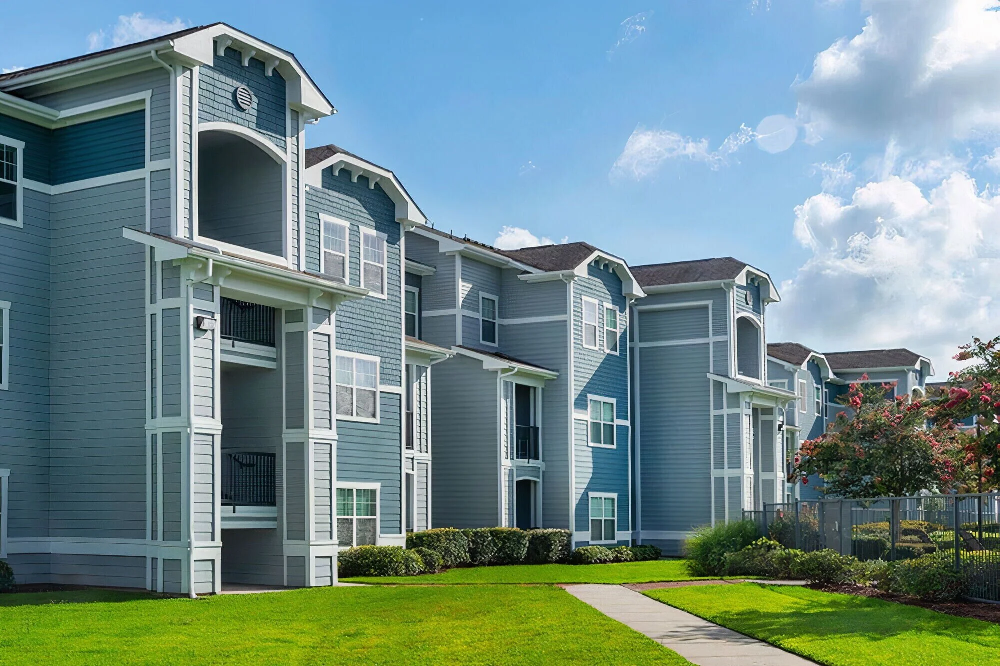 Multi-story residential apartment building painted in shades of blue with white trim, green lawn, walking path, garden bushes, and flowering trees under a partly cloudy sky.