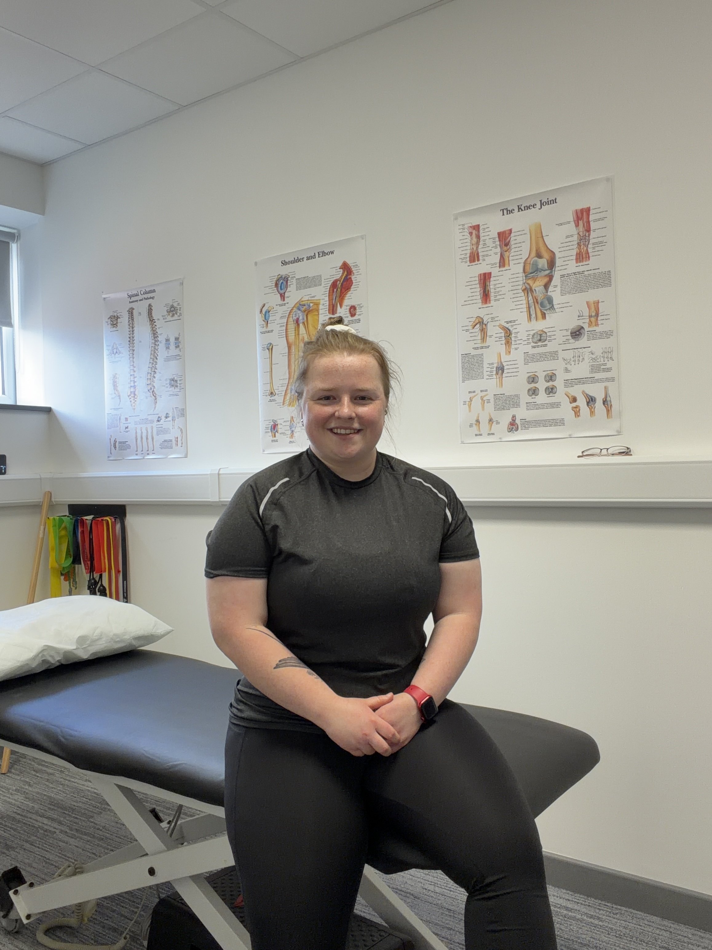 A woman in black athletic clothing sitting on an examination table in a medical office, with anatomical posters on the wall behind her.