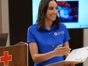 A woman in a blue shirt standing at a podium with a Red Cross logo, smiling and holding a pen.