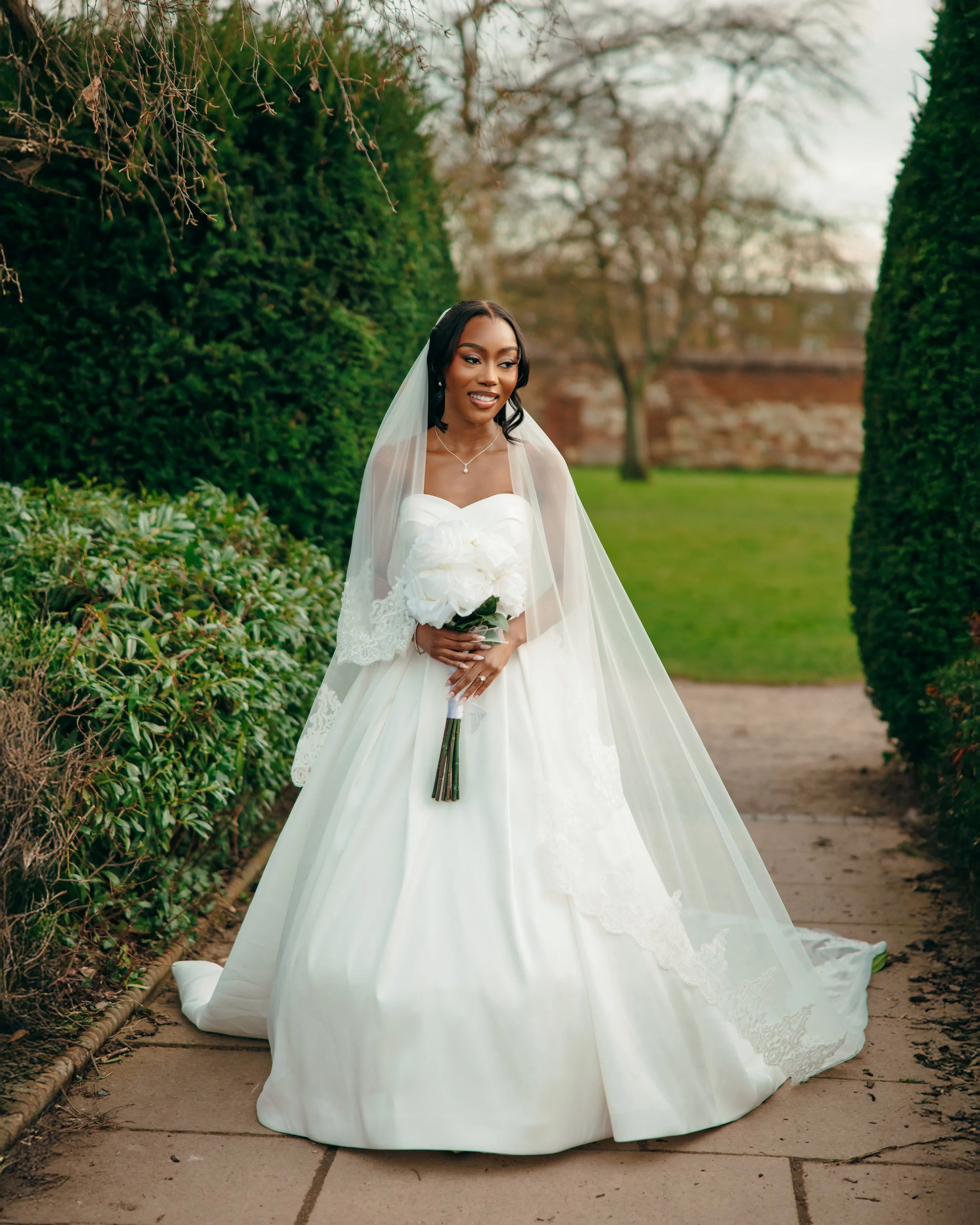 Bride in a white wedding gown with lace details, holding a bouquet of white roses, standing on a garden pathway surrounded by greenery and trees.