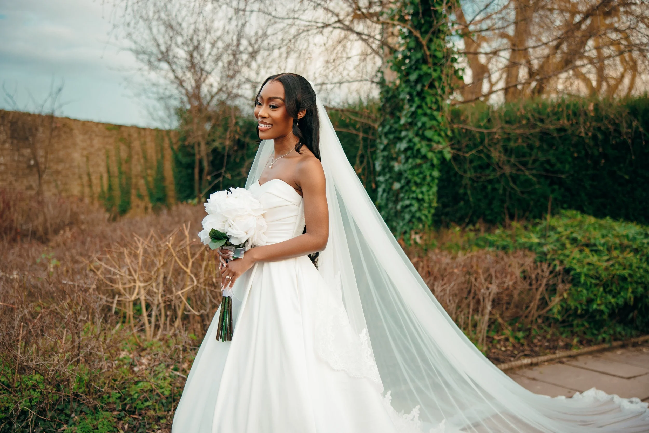 A smiling bride in a white wedding gown holding a bouquet of white flowers outdoors, with trees and bushes in the background.