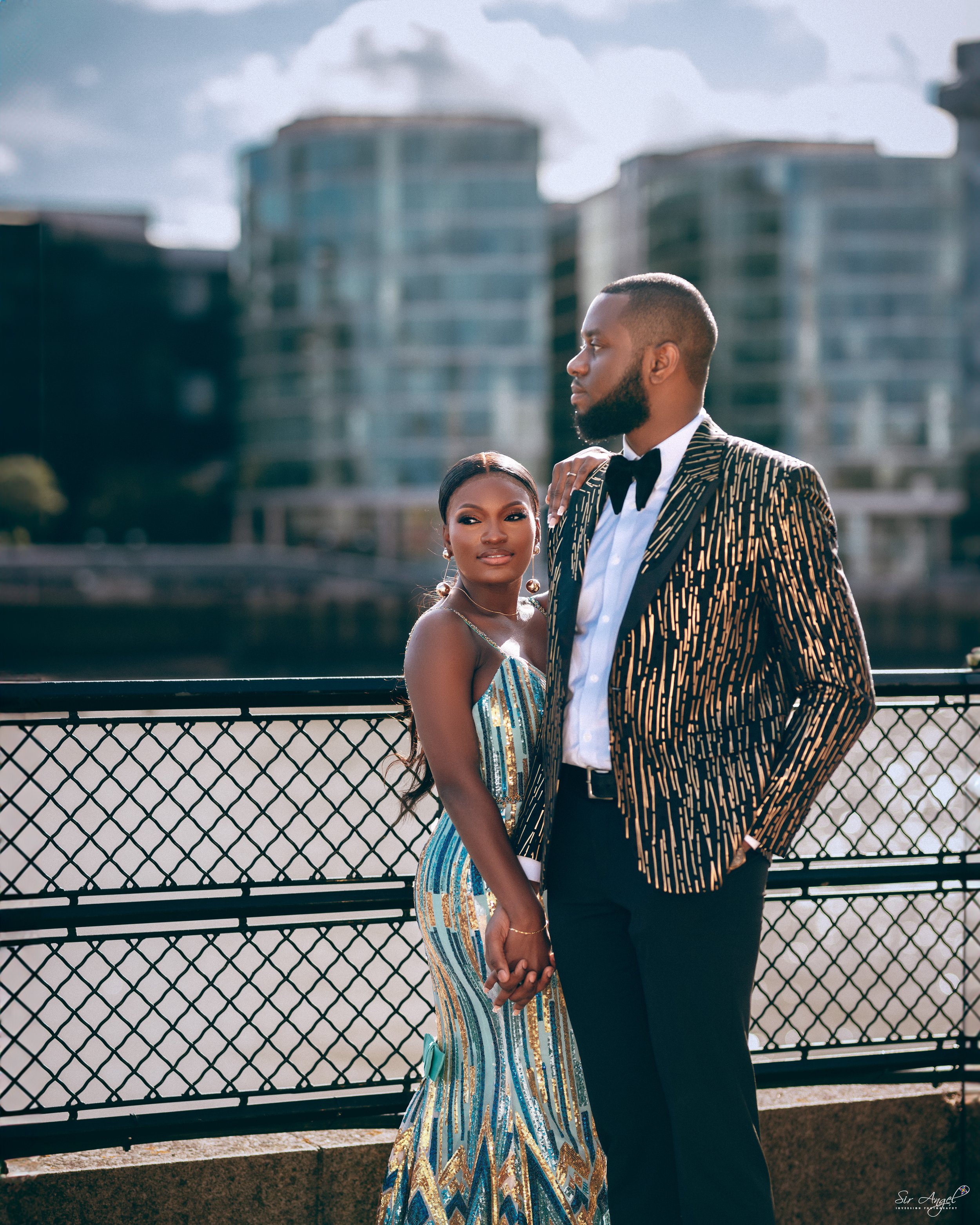 A stylishly dressed couple holding hands by a river, with modern buildings in the background, during daytime.
