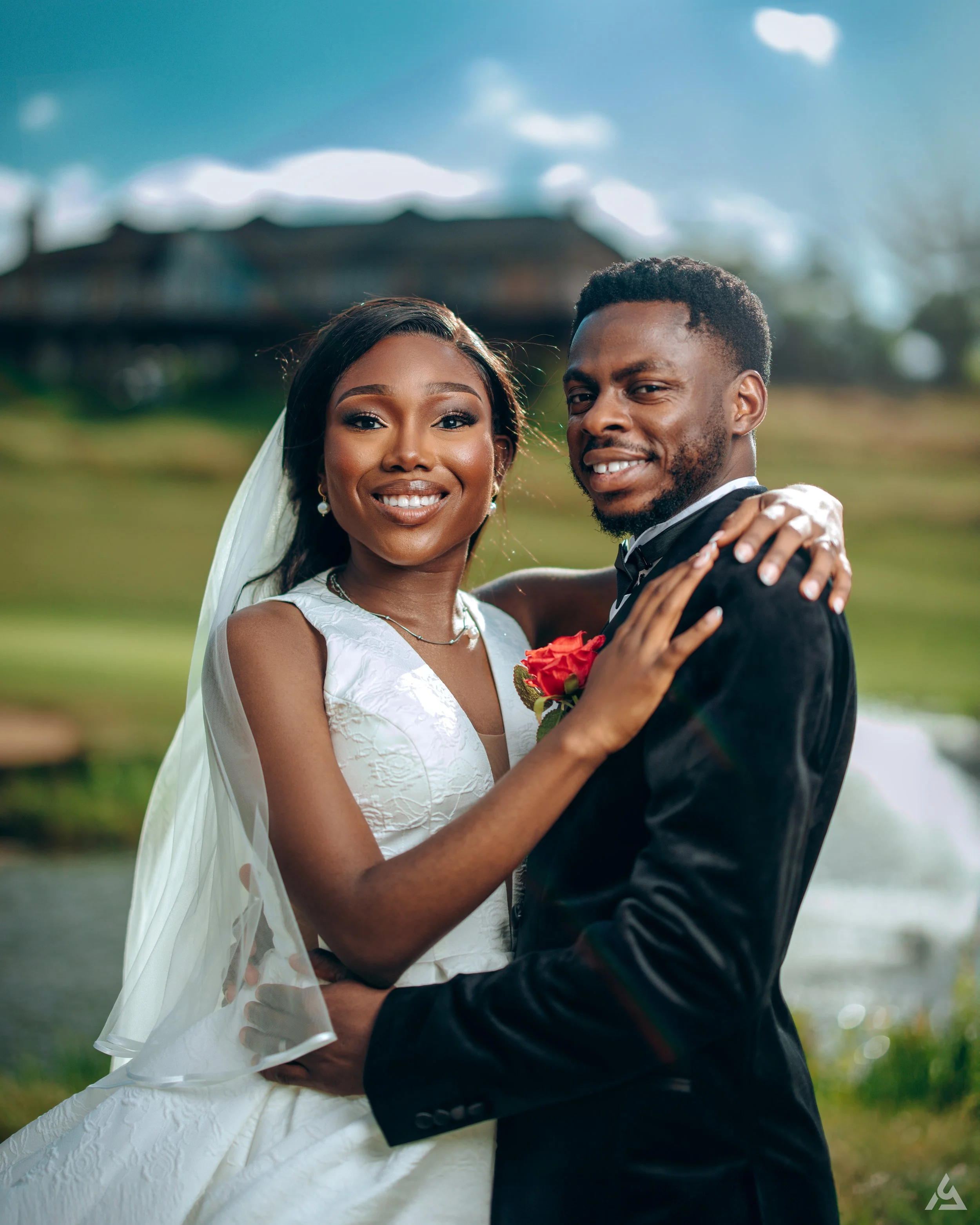A smiling couple in wedding attire standing outdoors by a pond, with a house and cloudy sky in the background.