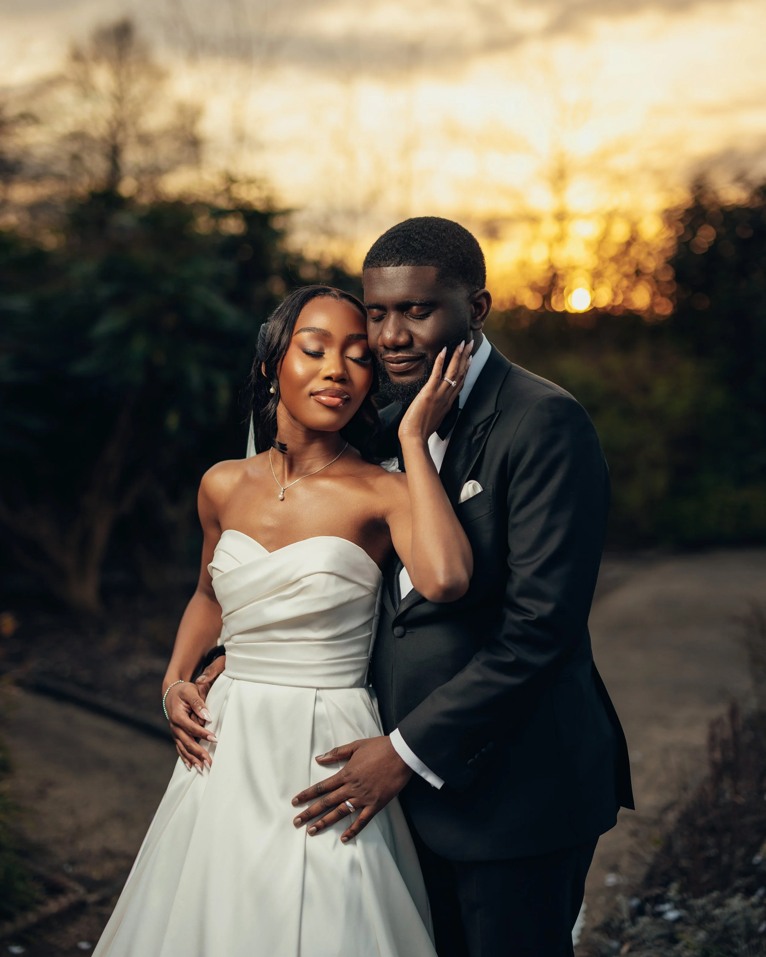 A bride and groom in wedding attire sharing an intimate moment outdoors at sunset, with the bride touching the groom's face and both closing their eyes.