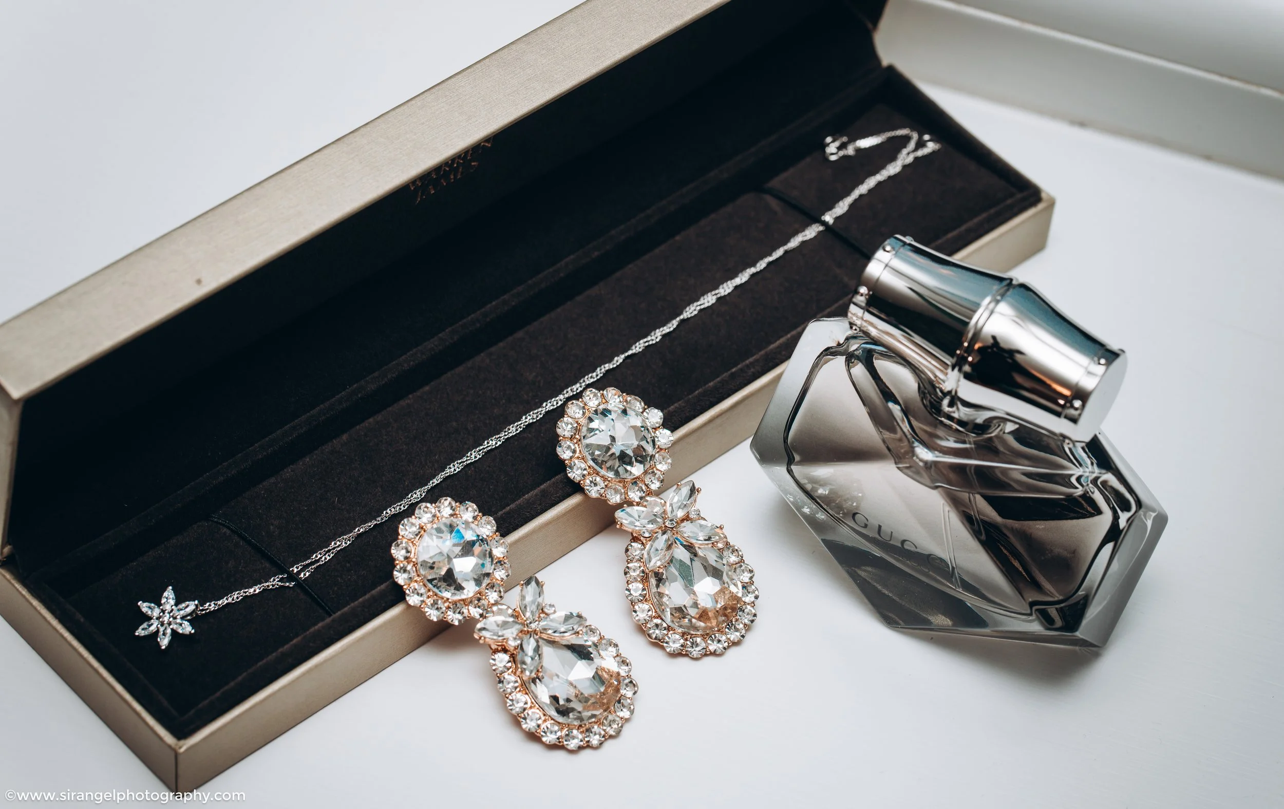 A jewelry box with a necklace and earrings, a bottle of Gucci perfume, and a metallic flashlight on a white surface.