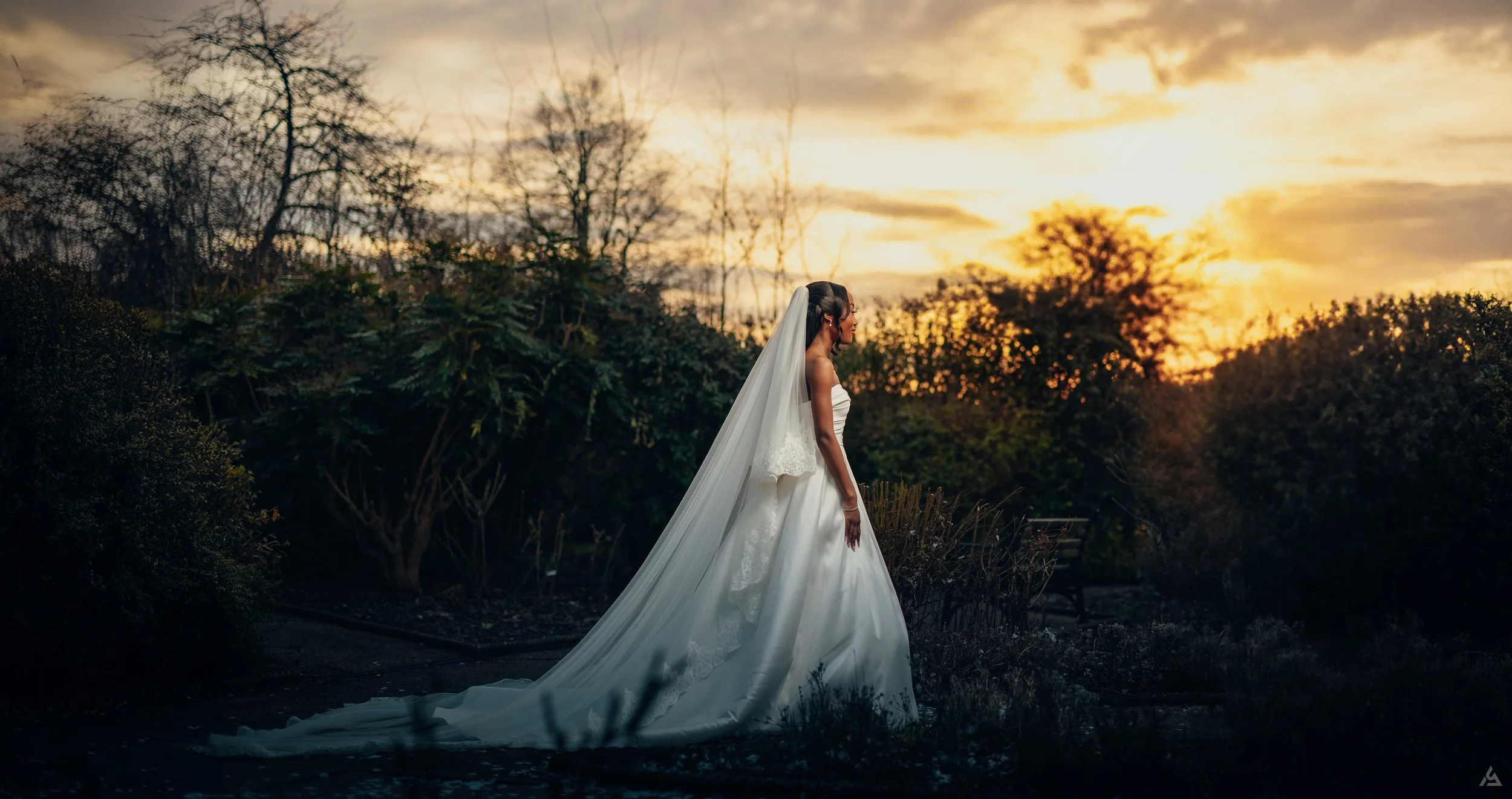 A bride in a white wedding gown with a long train and veil stands outdoors at sunset, surrounded by trees and bushes.