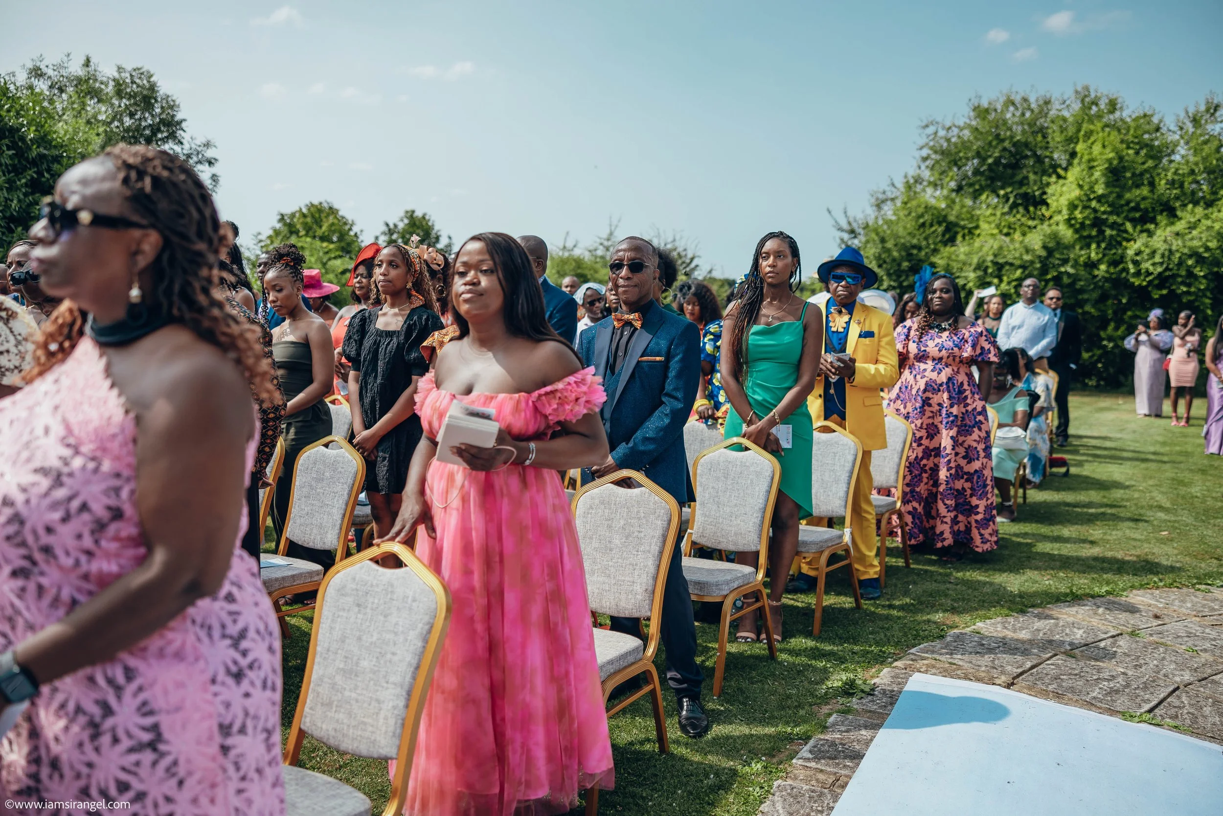 People attending an outdoor event, standing and seated, dressed in formal and colorful attire on a grassy area under a clear sky.