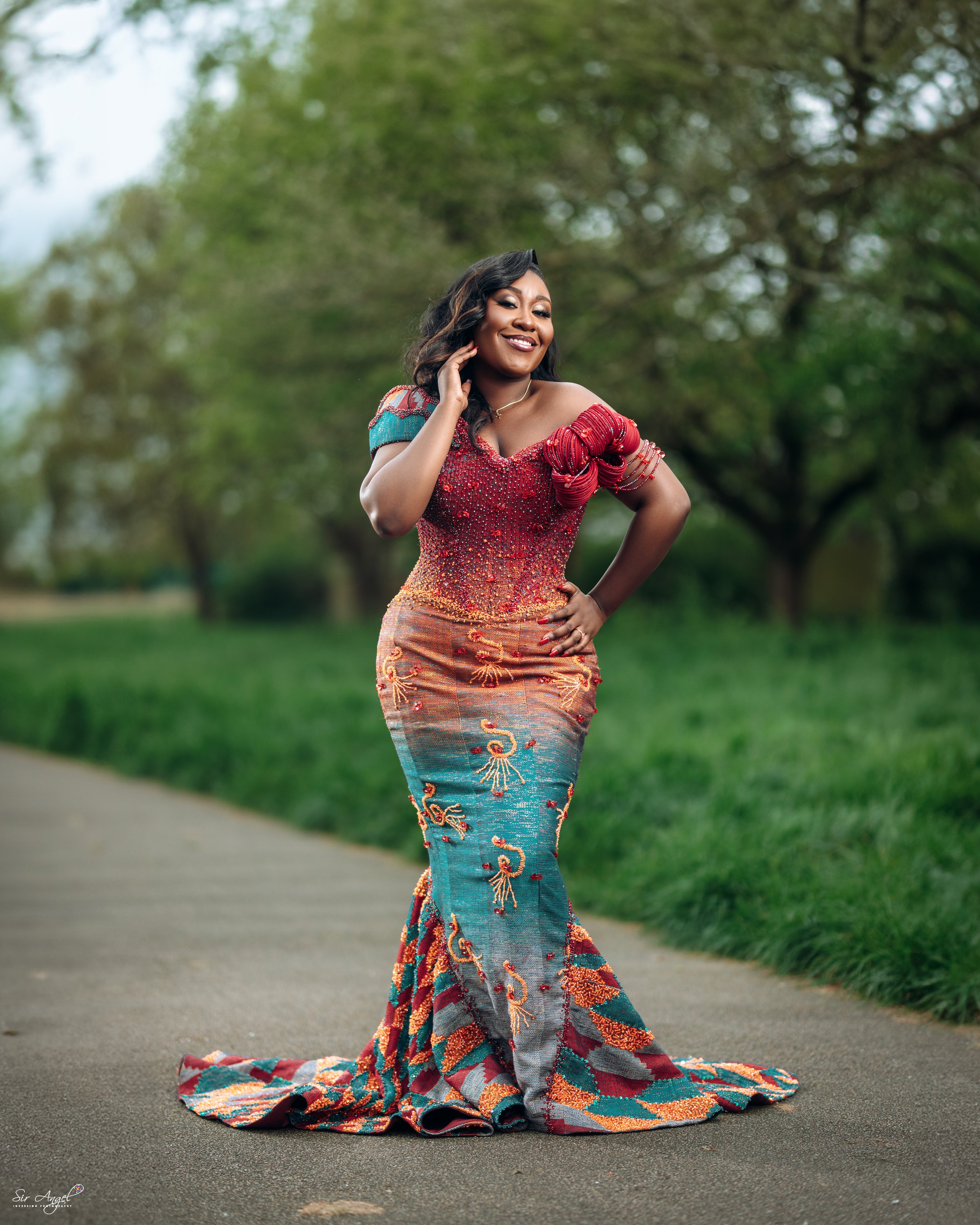 Woman in a colorful, traditional gown posing outdoors on a paved path with green trees in the background.