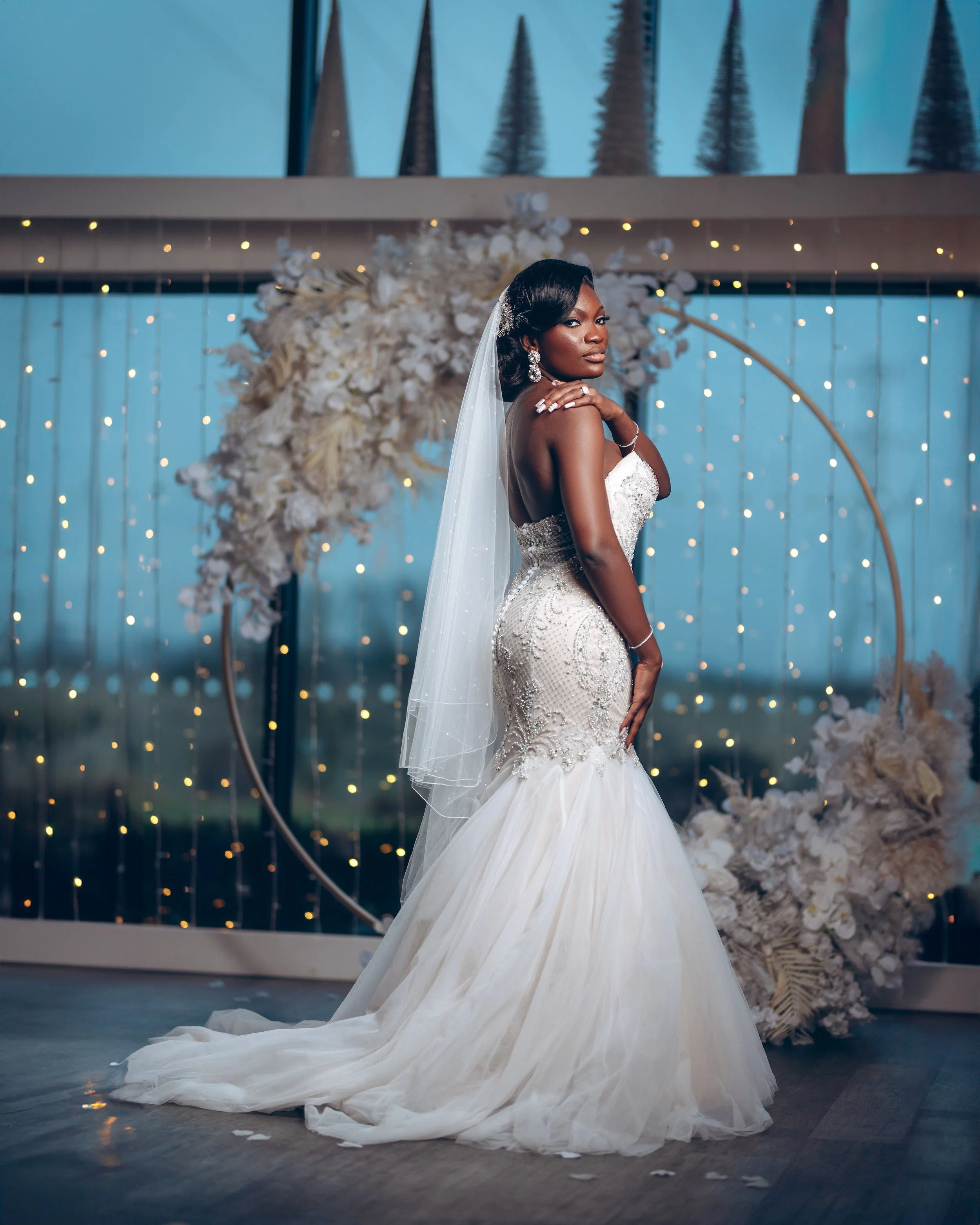 A bride in a white wedding gown with lace details, standing in front of a floral backdrop with fairy lights, on a wooden floor, posing with one hand on her shoulder and the other on her waist.