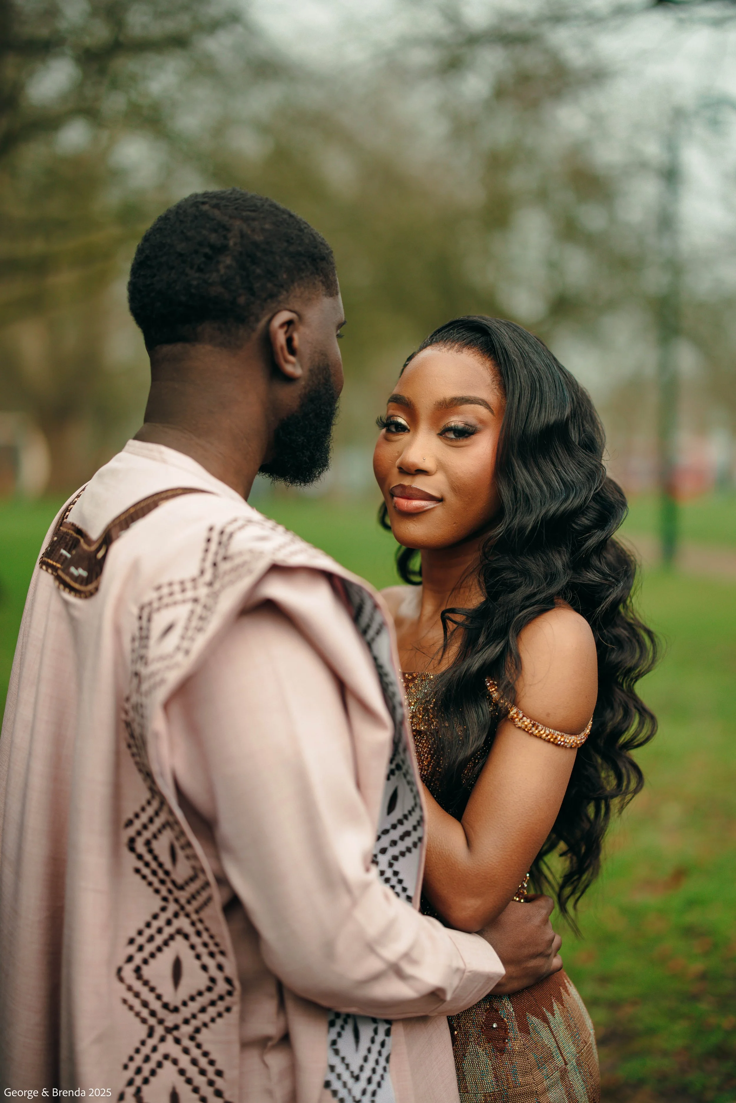 A couple standing close together outdoors, with the man facing away and the woman looking directly at the camera, in a park with green grass and blurred trees in the background.
