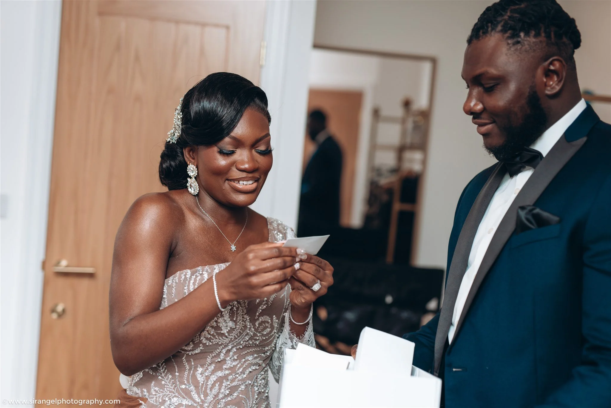 A bride and groom exchanging gifts at a wedding, both dressed in formal attire, inside a home with a wooden door and interior decor.