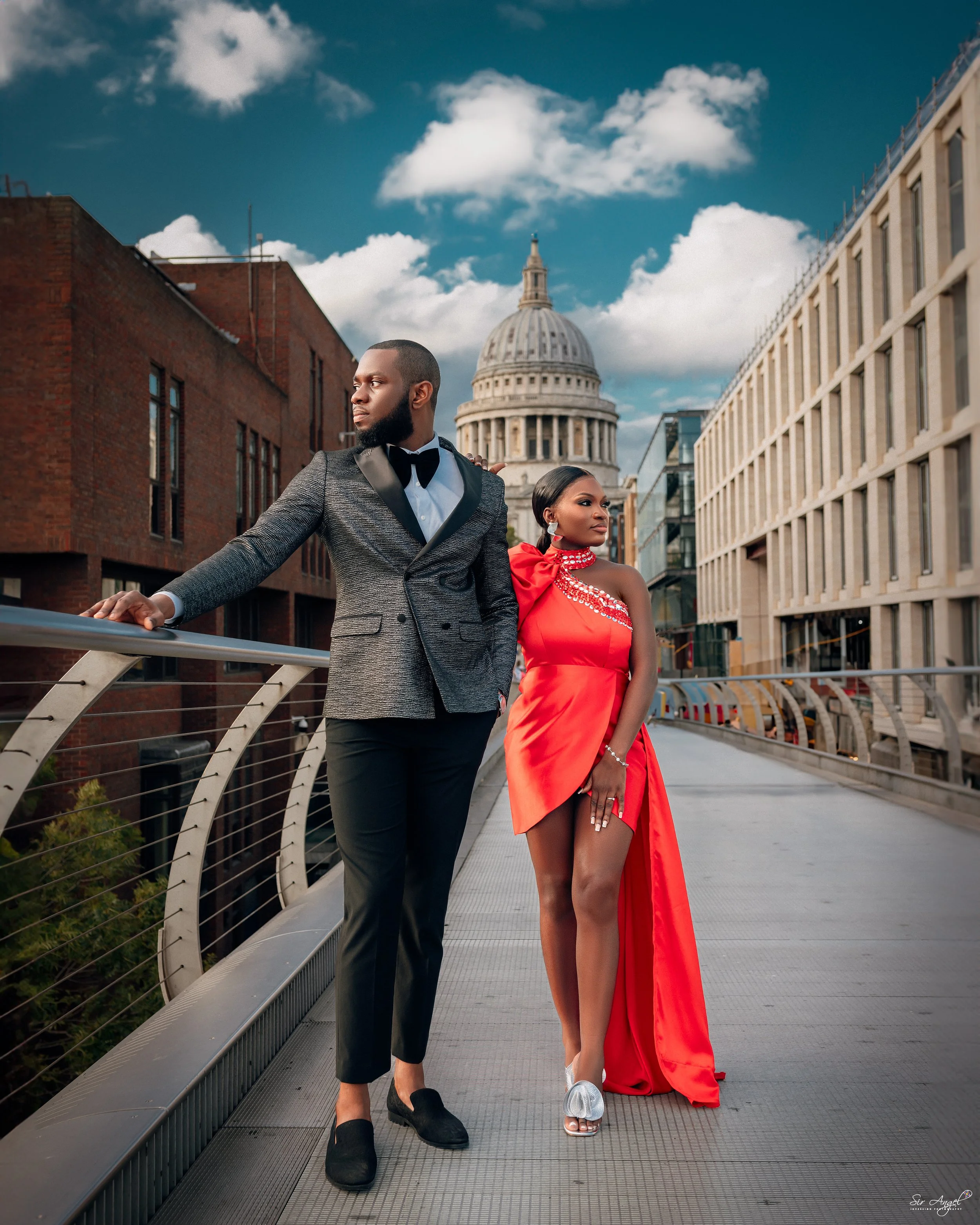 A stylish man in a tuxedo and a woman in a red evening gown walk on a city bridge with the United States Capitol in the background, under a partly cloudy sky.