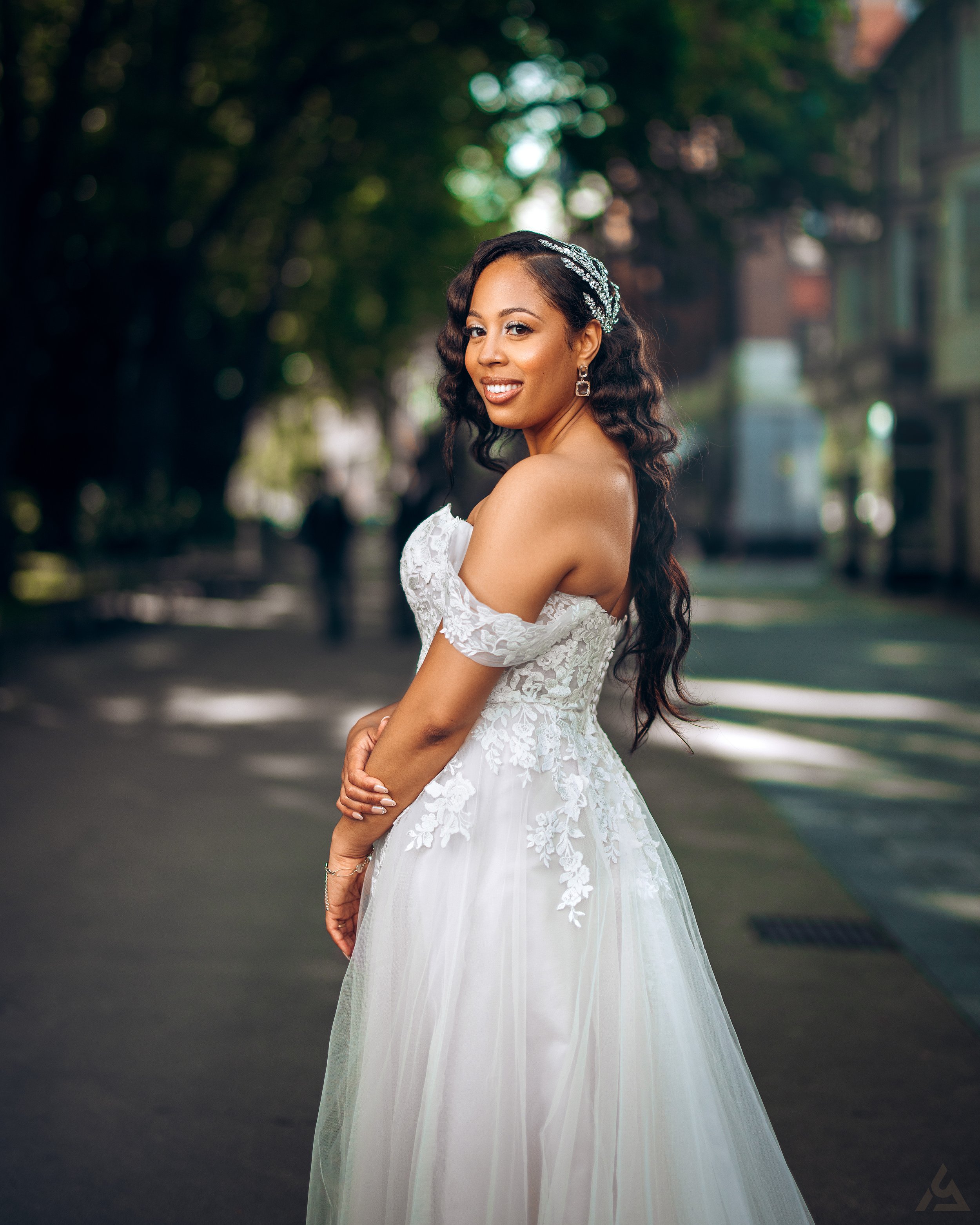 A woman in a white wedding dress smiling and posing outdoors on a shaded street.
