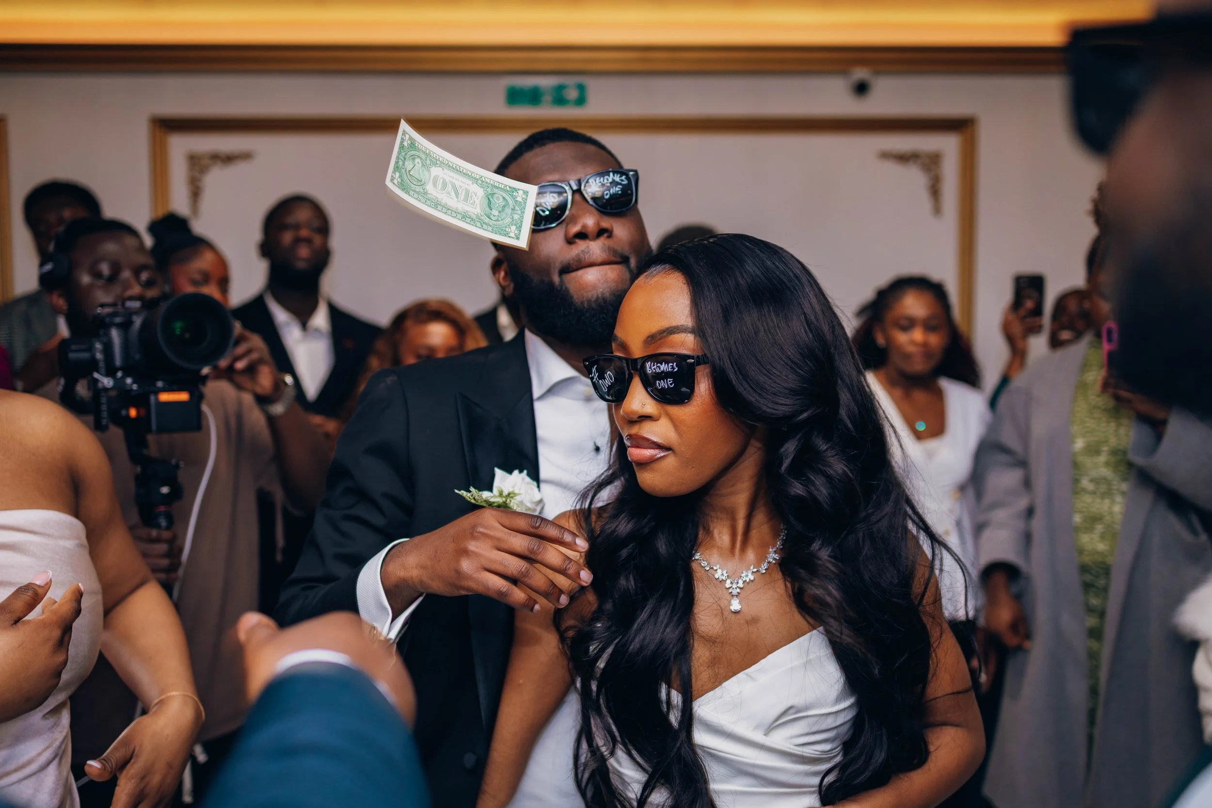 A couple at a wedding or celebration wearing matching sunglasses with the words 'Groom' and 'Bride' written on them. The man is wearing a suit with a boutonniere and has a dollar bill hanging from his sunglasses. The woman is dressed in a white dress