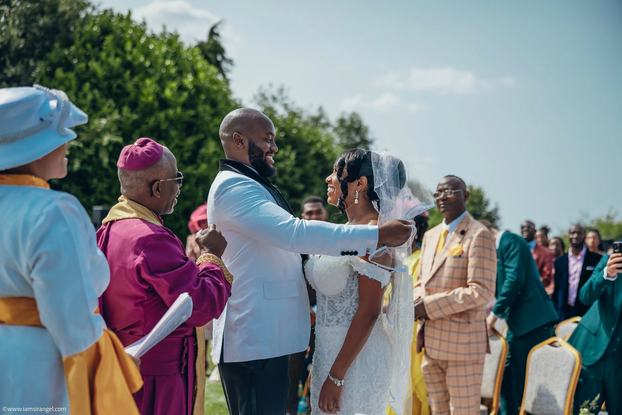 A wedding ceremony outdoors featuring a bride and groom smiling at each other, surrounded by friends and family, with green trees and a blue sky in the background.