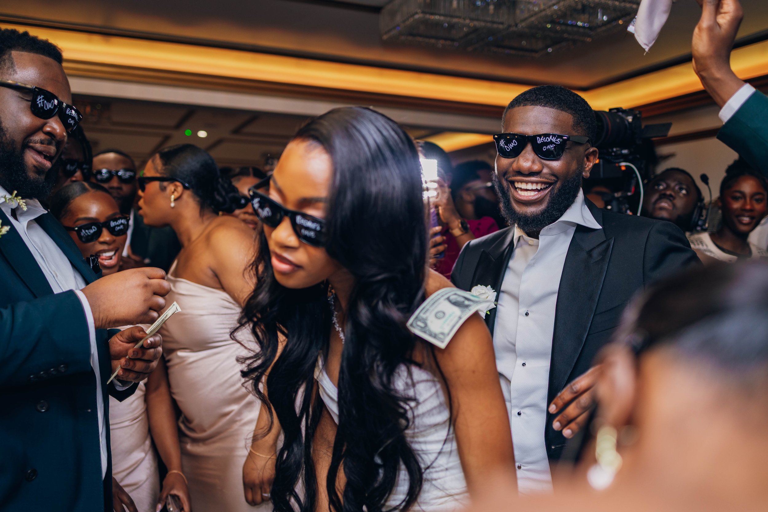 People celebrating at a wedding reception, wearing dark glasses and smiling, with money tucked into the bride's dress.