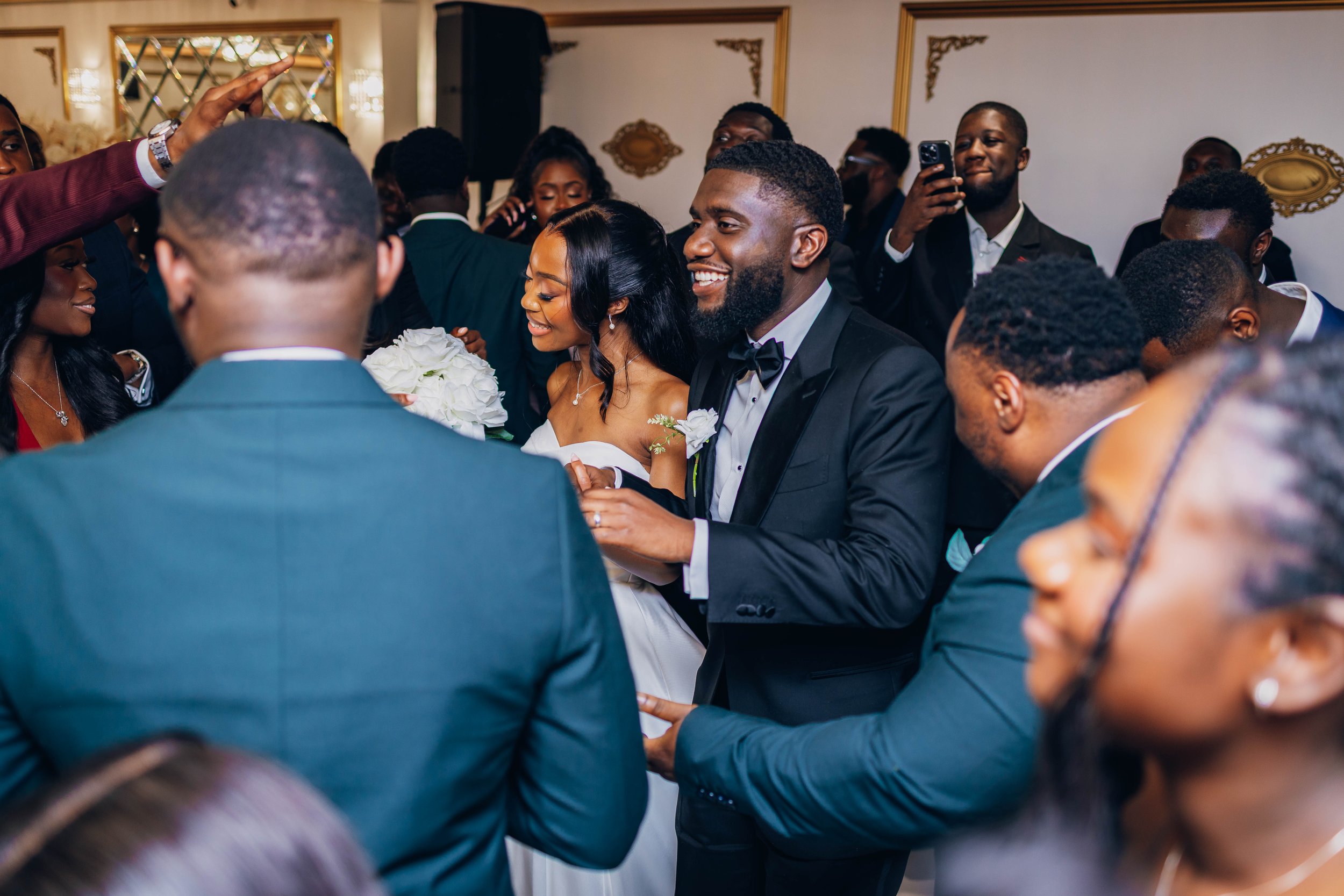 A wedding reception with a bride and groom smiling and holding hands, surrounded by guests in formal attire, some taking photos and videos, in a decorated indoor venue.