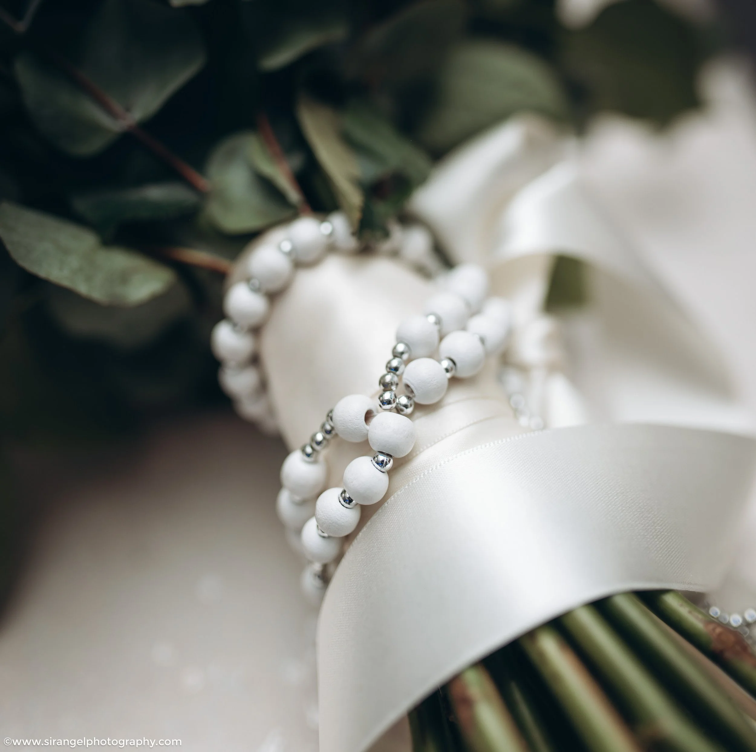 Close-up of a bridal bouquet wrapped with white satin ribbon, decorated with pearl and silver bead jewelry, with green leaves in the background.