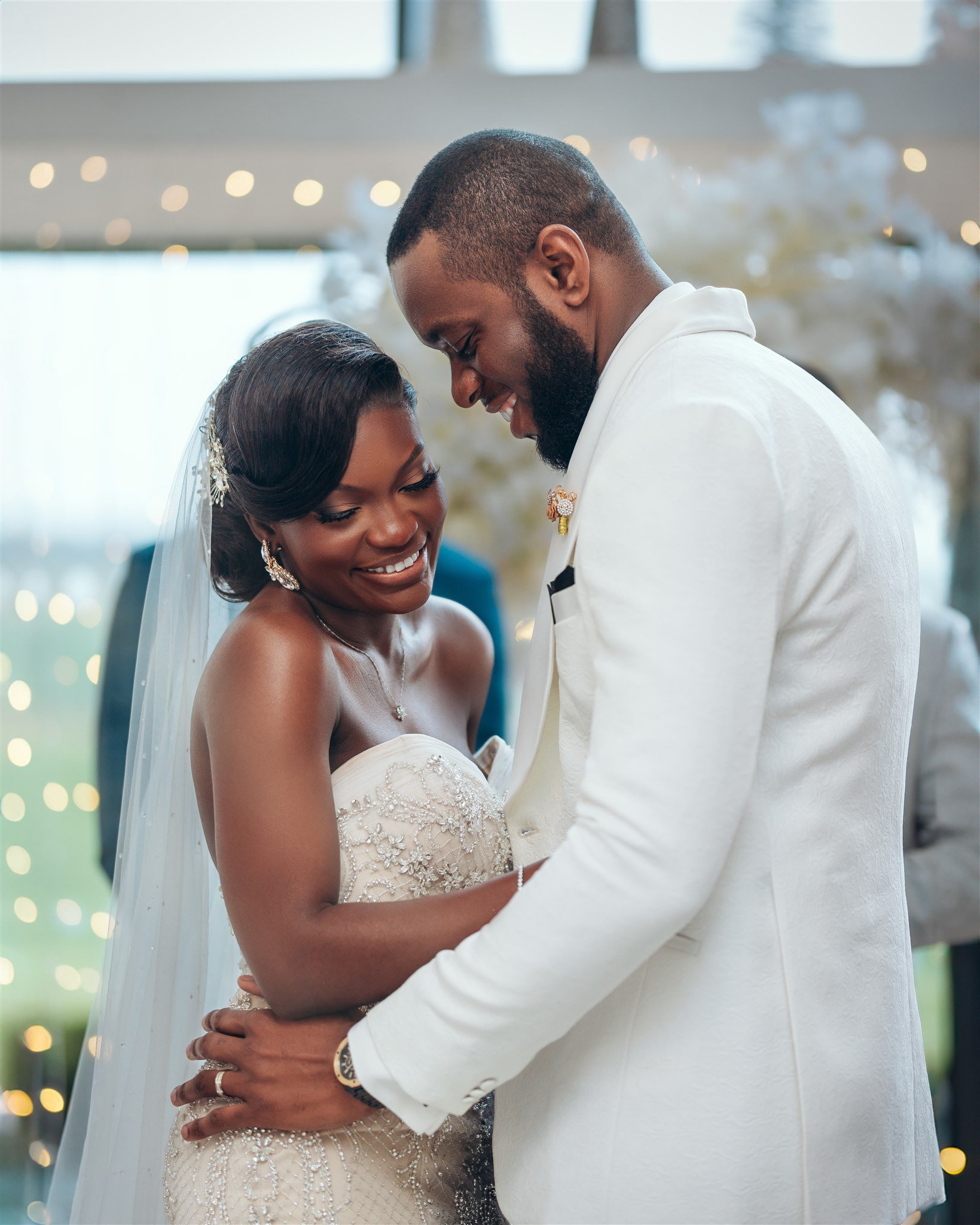 A bride and groom in wedding attire share an intimate moment, smiling and holding each other closely in a decorated indoor venue.