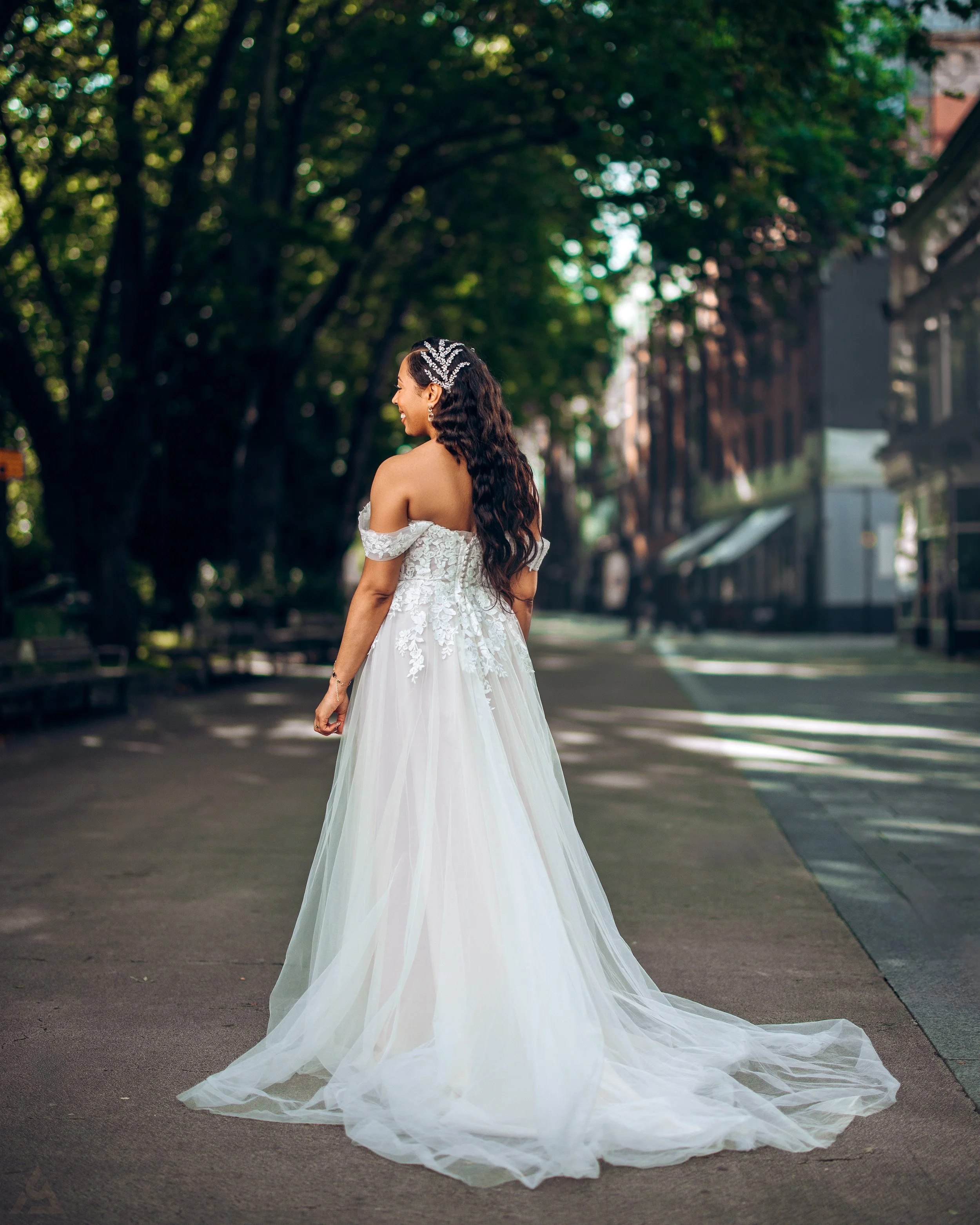 A woman in a white wedding dress with lace details and off-the-shoulder sleeves, standing on a city street with trees and buildings in the background. She has long dark hair with a decorative headpiece and is turning slightly to the side, smiling.