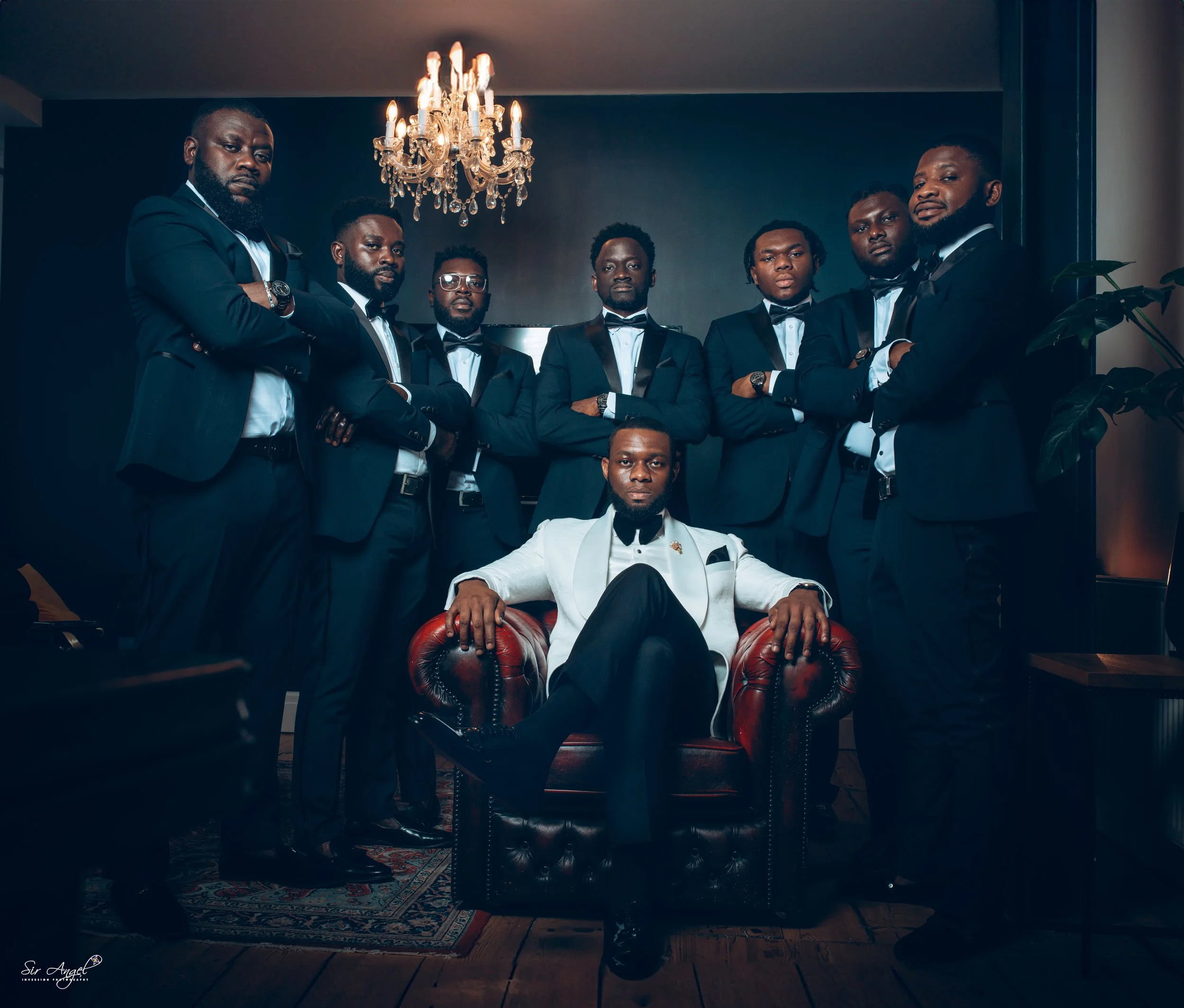 A group of nine men dressed in black tuxedos, with one man seated in a white tuxedo sitting on a red leather armchair in front of a black background. The men stand with arms crossed or at their sides under a chandelier.