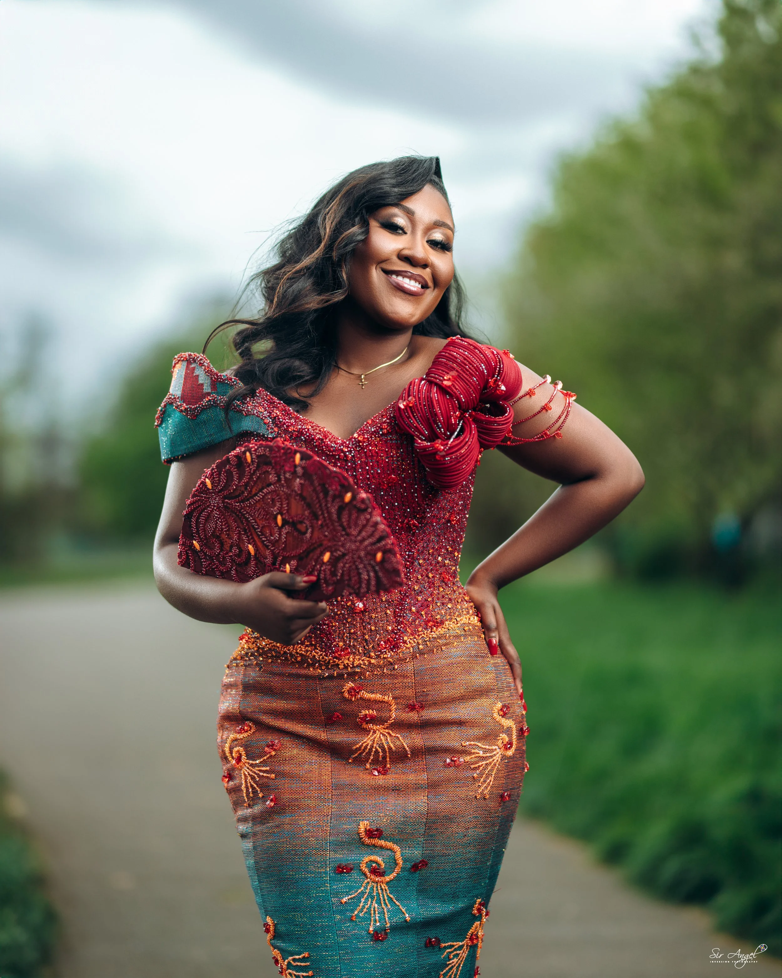 A woman wearing a colorful, traditional African dress with beaded embroidery and holding a matching headwrap, smiling outdoors on a cloudy day.