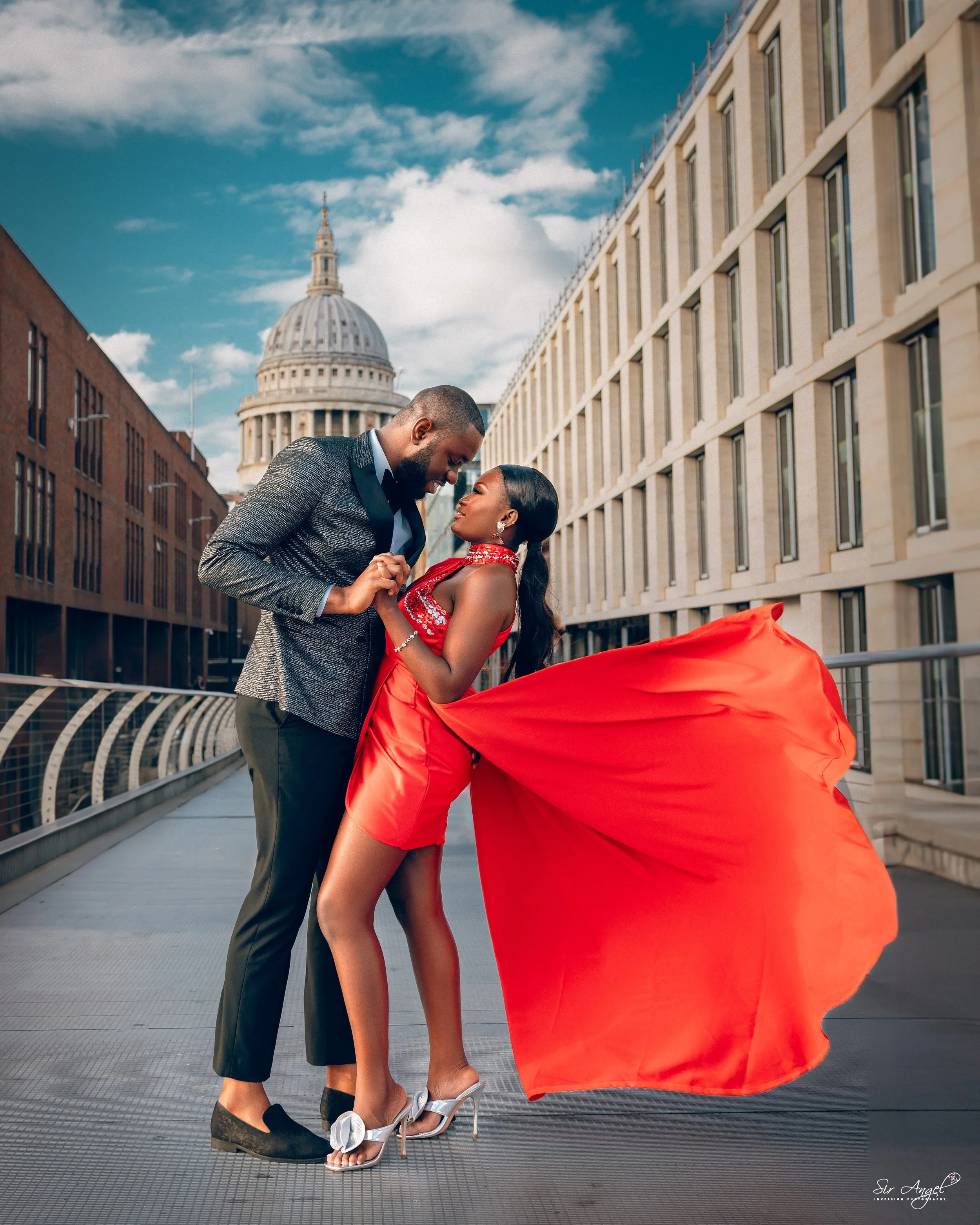 A couple standing close on an urban bridge with buildings and a domed government building in the background, holding hands and looking at each other, with the woman wearing a flowing red dress and high heels.