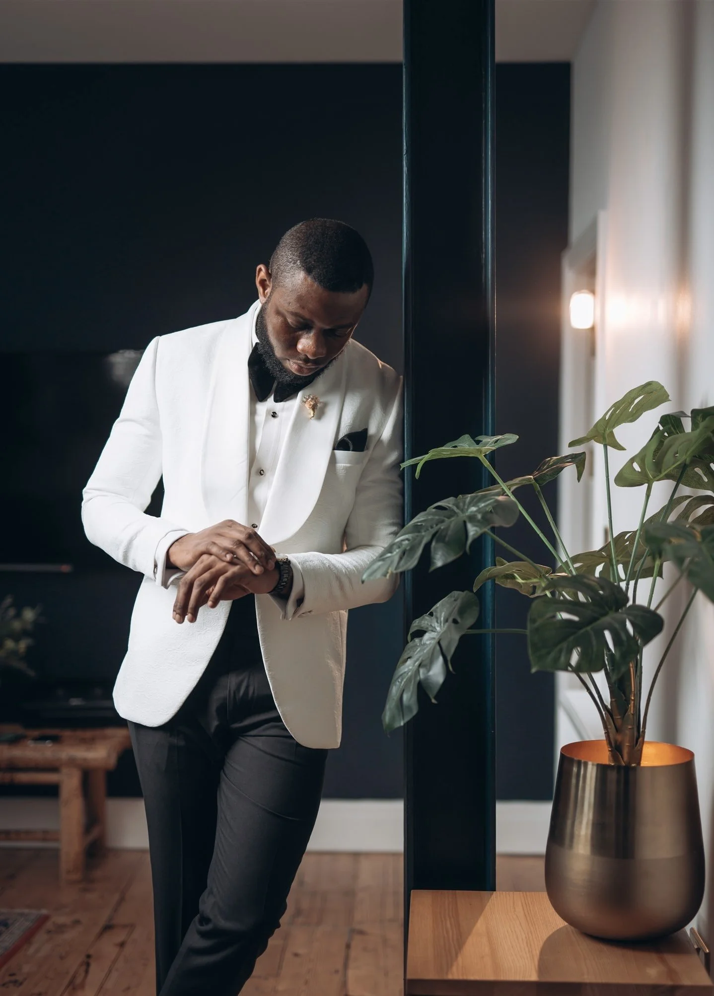 A man dressed in a white tuxedo jacket with black pants, bow tie, and pocket square, standing indoors and leaning against a wall with greenery in a gold planter.