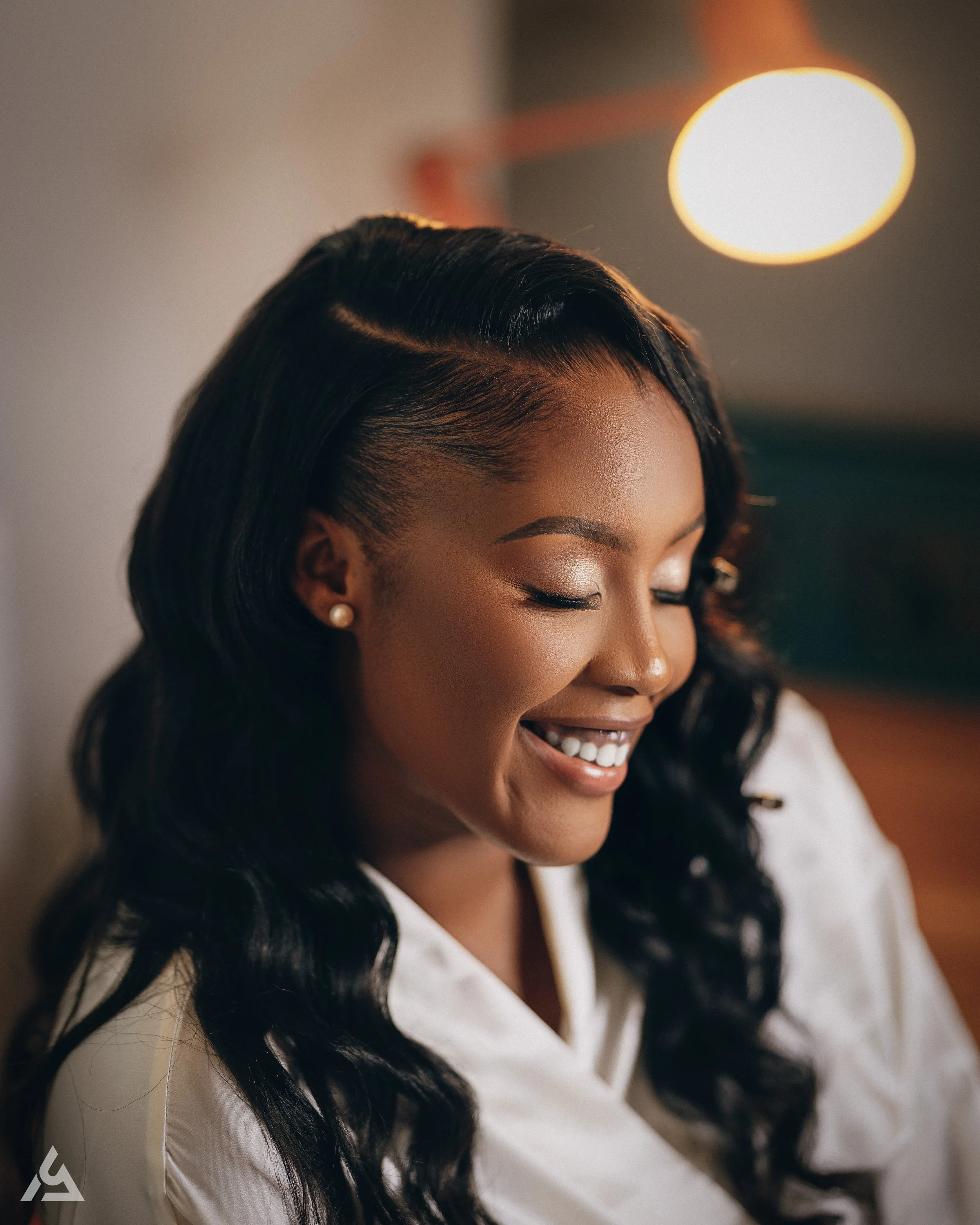 Close-up of a smiling African American woman with long, wavy black hair, wearing pearl earrings and a white blouse, with warm lighting and a blurred background.