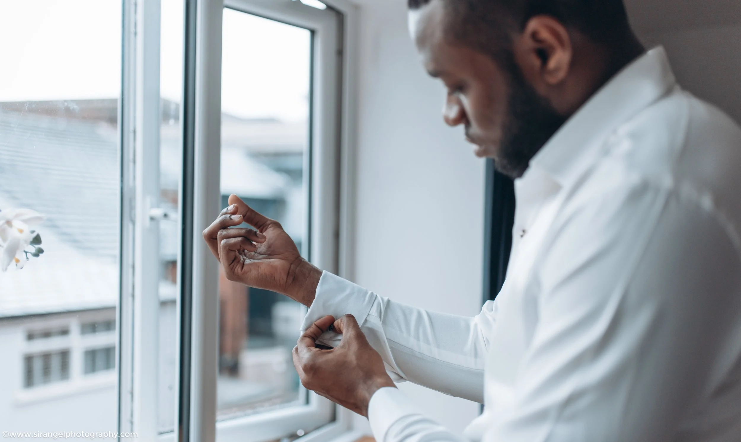 A man in a white shirt adjusts his cuff near a window with a view of neighboring buildings.