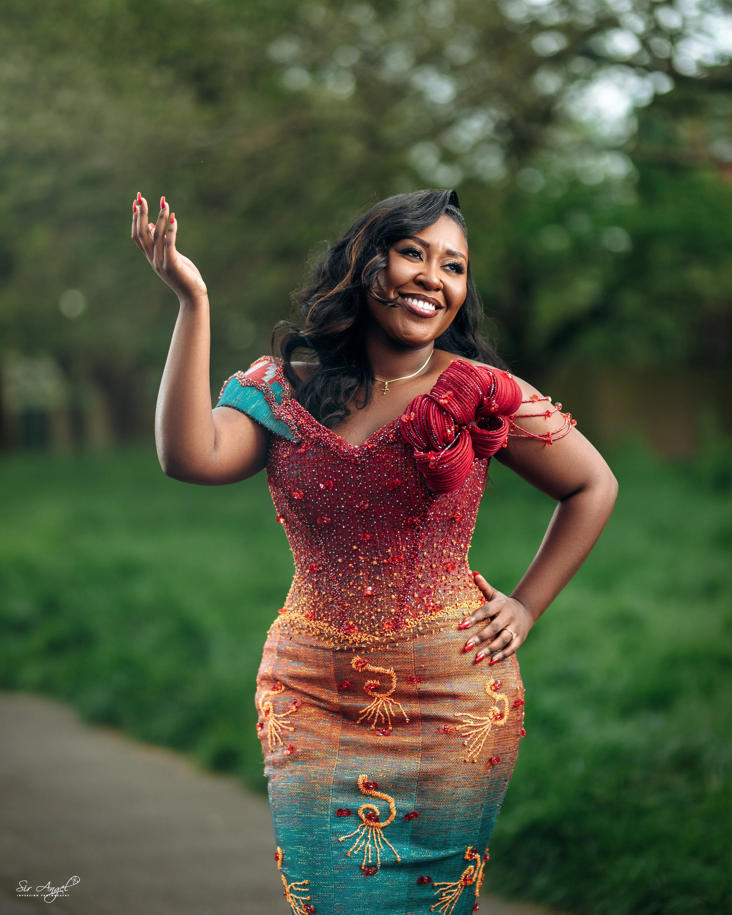 A woman smiling and waving outdoors, wearing an elegant, colorful dress with intricate embroidery and beadwork, set against a green, blurry background.