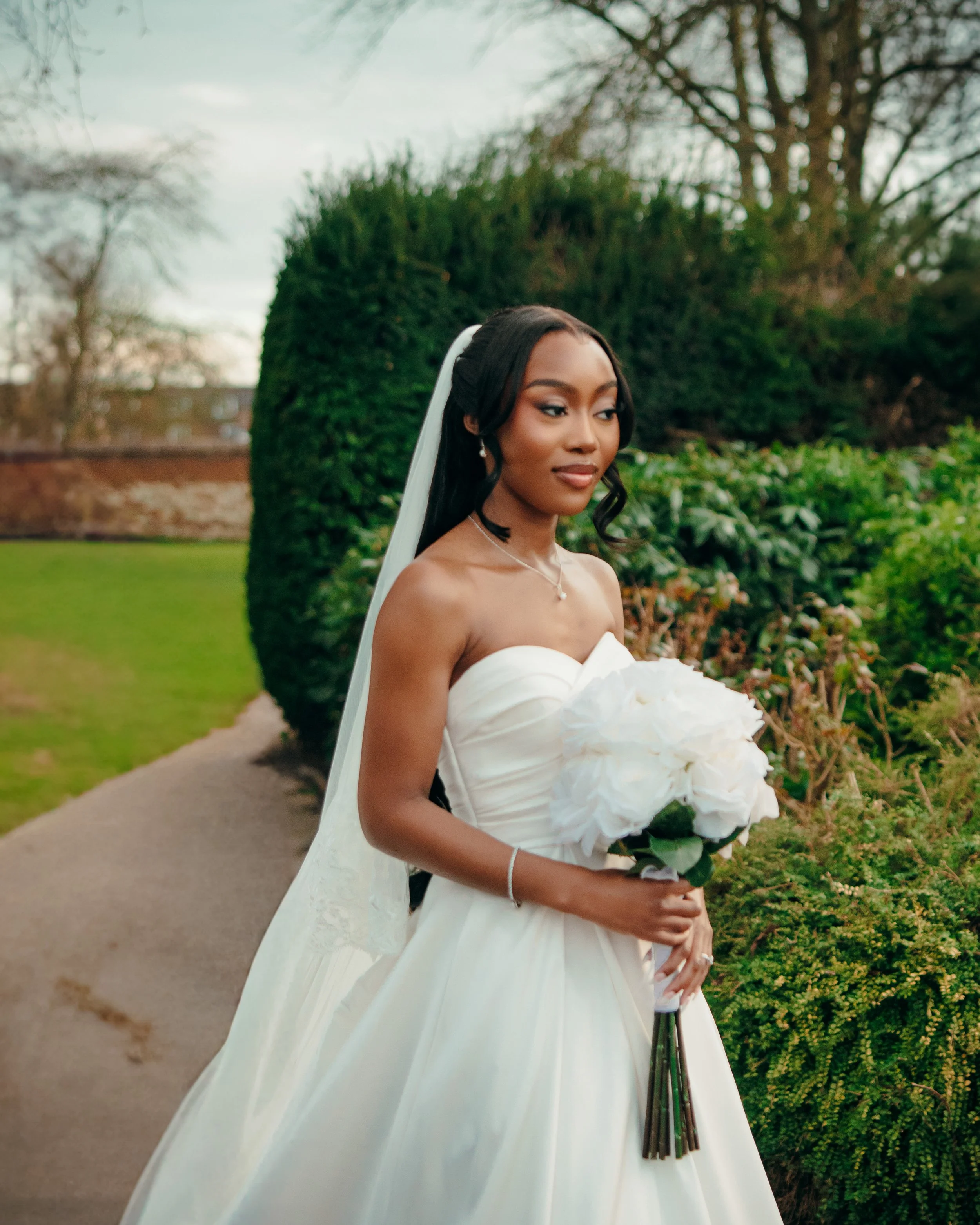 A bride in a white wedding dress holding a bouquet of white flowers standing outdoors in a garden.