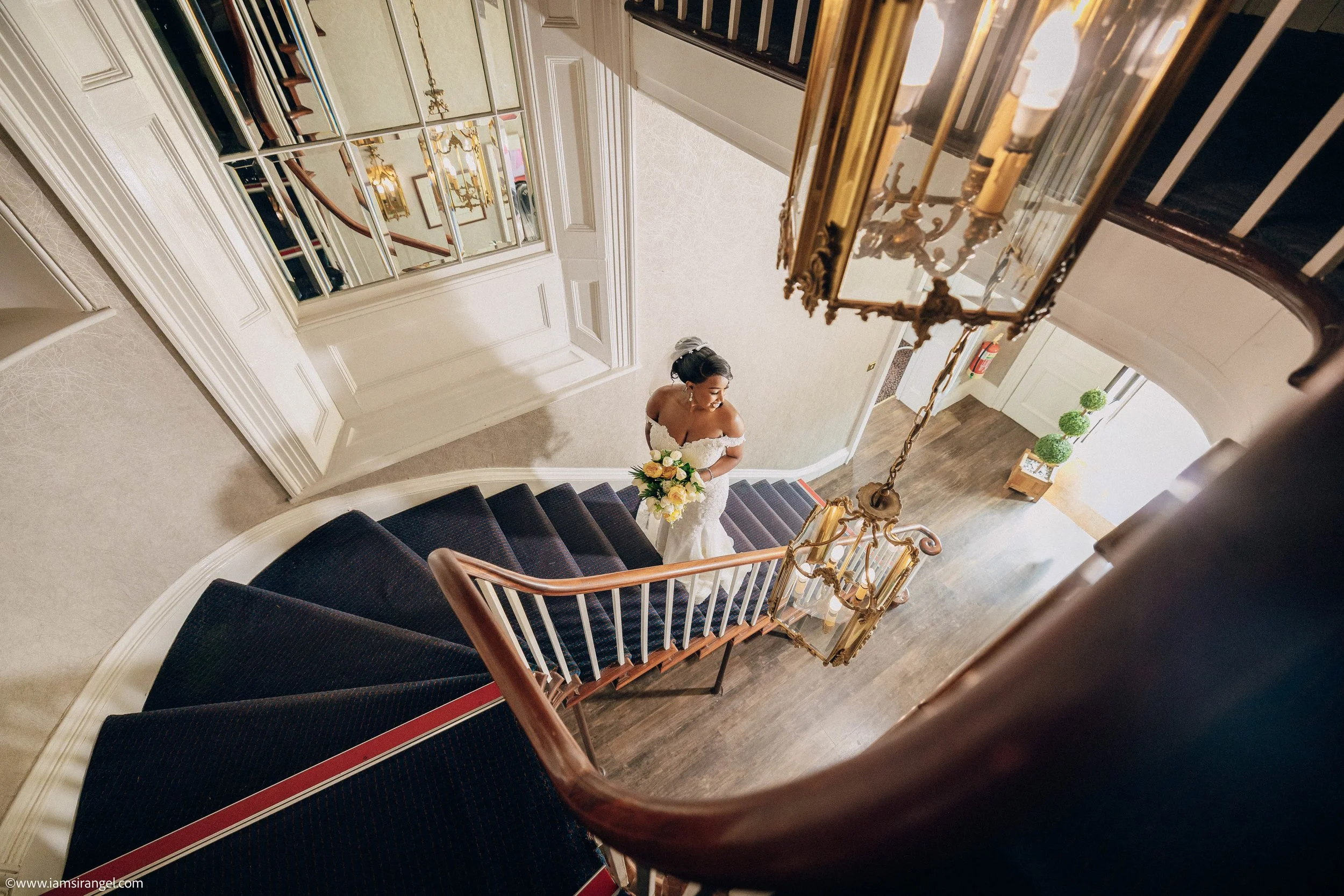 A bride in a white wedding dress holding a bouquet of yellow and white flowers standing on a staircase in a fancy interior with mirrors and chandeliers.