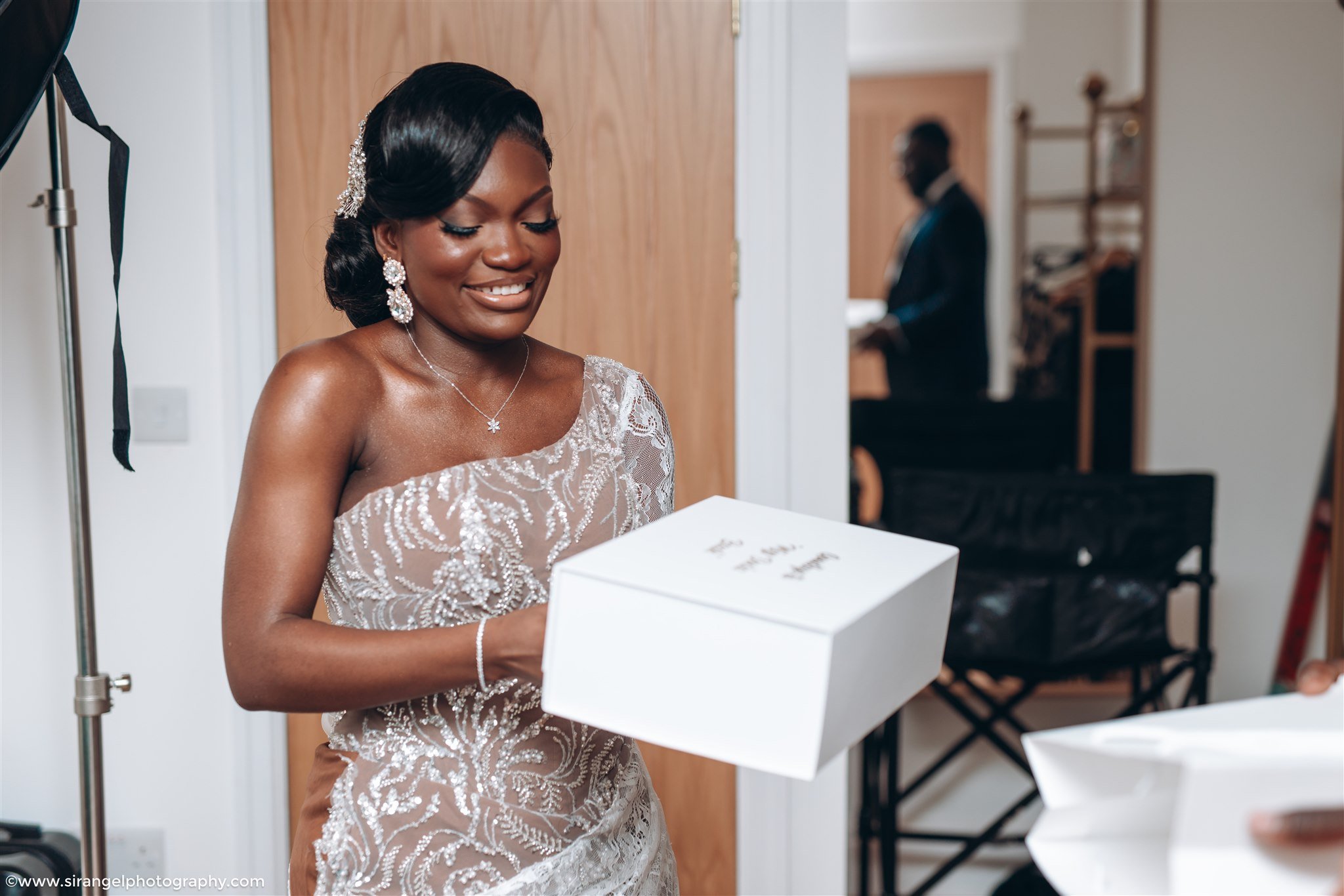 A woman dressed in an elegant, beaded gown is smiling while opening a white gift box in a room, with a man in a suit playing piano in the background.