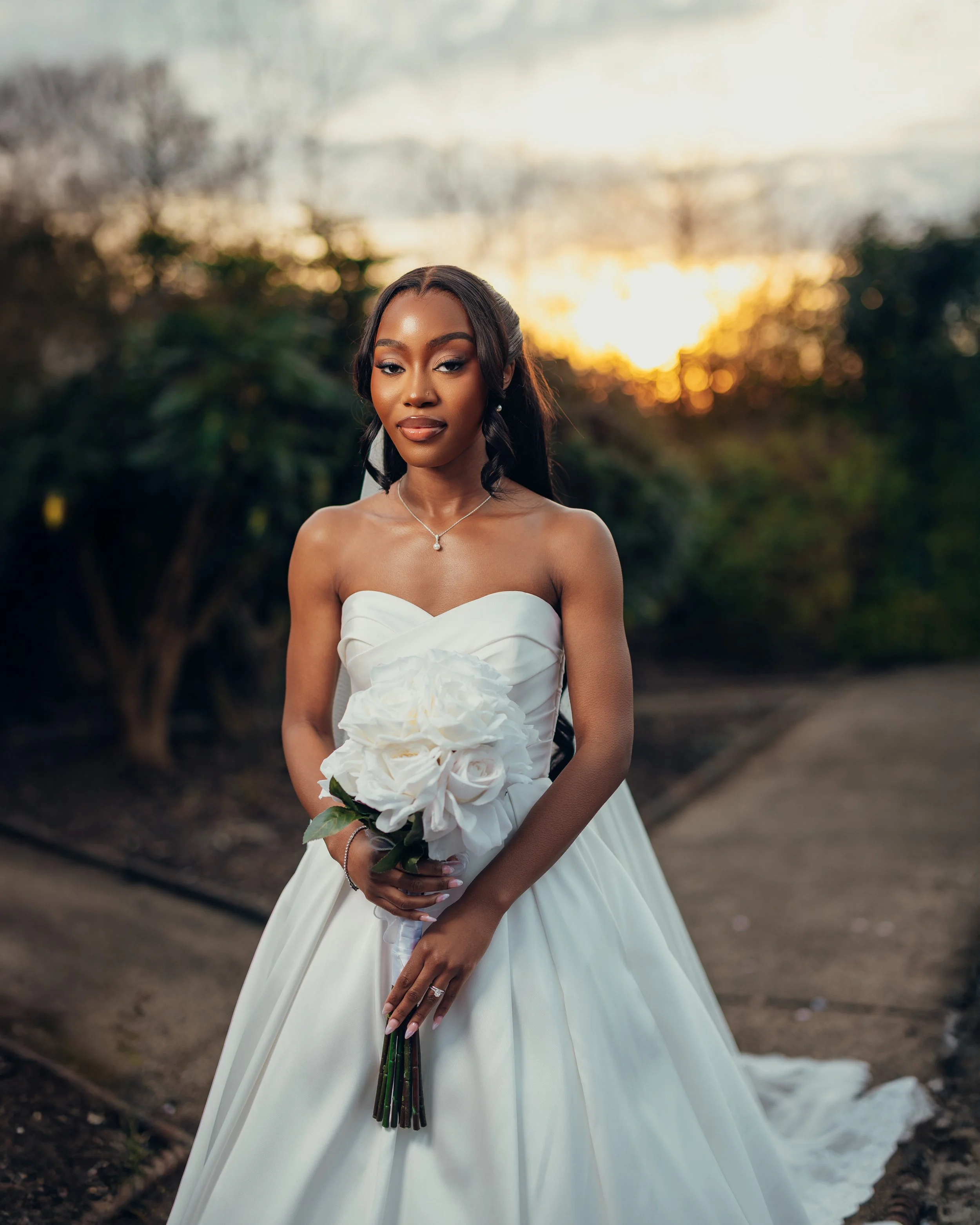 A bride in a strapless white wedding gown holding a bouquet of white roses and greenery outdoors during sunset.