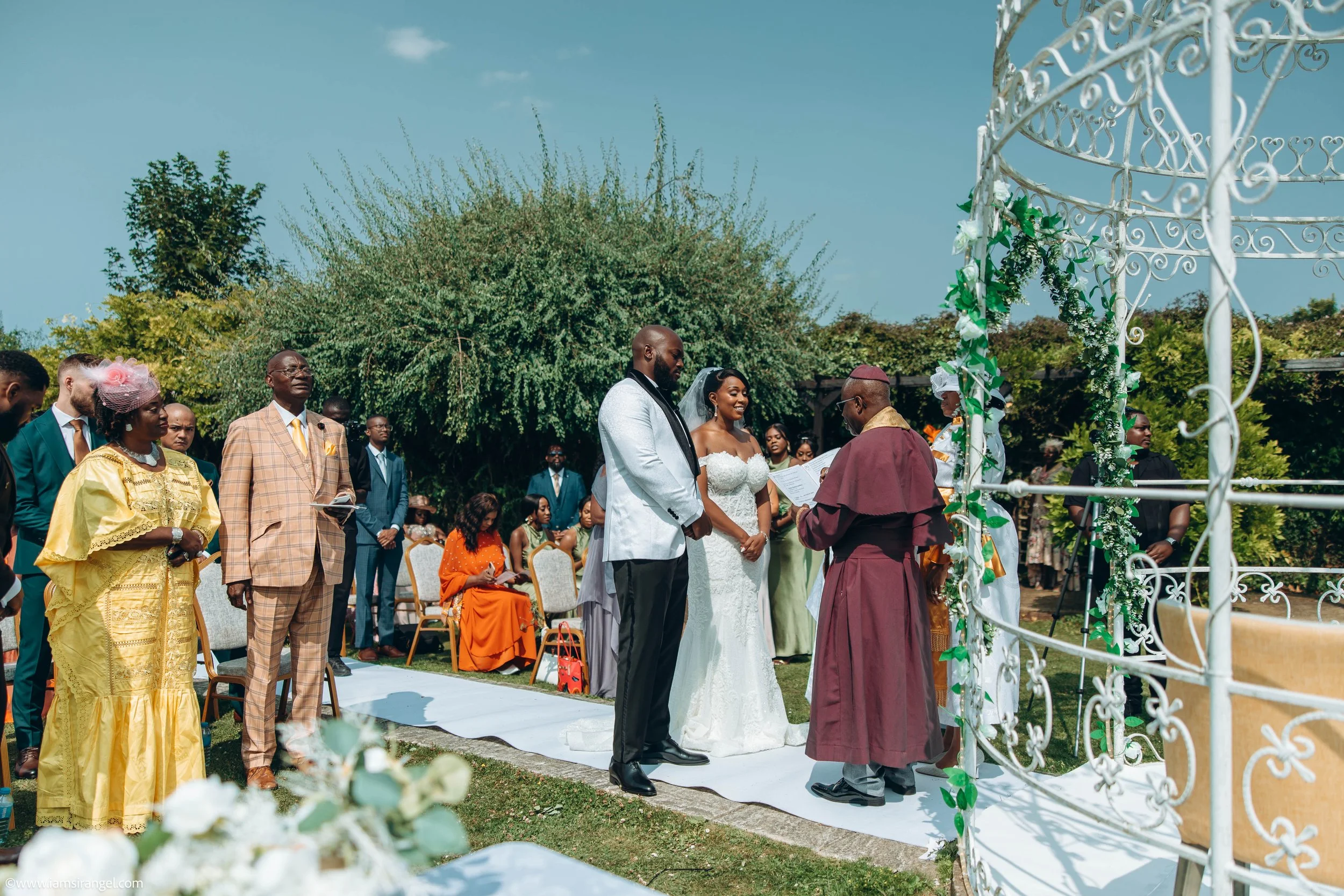A couple getting married at an outdoor wedding ceremony with guests seated around them, officiant standing in front, and natural greenery in the background.