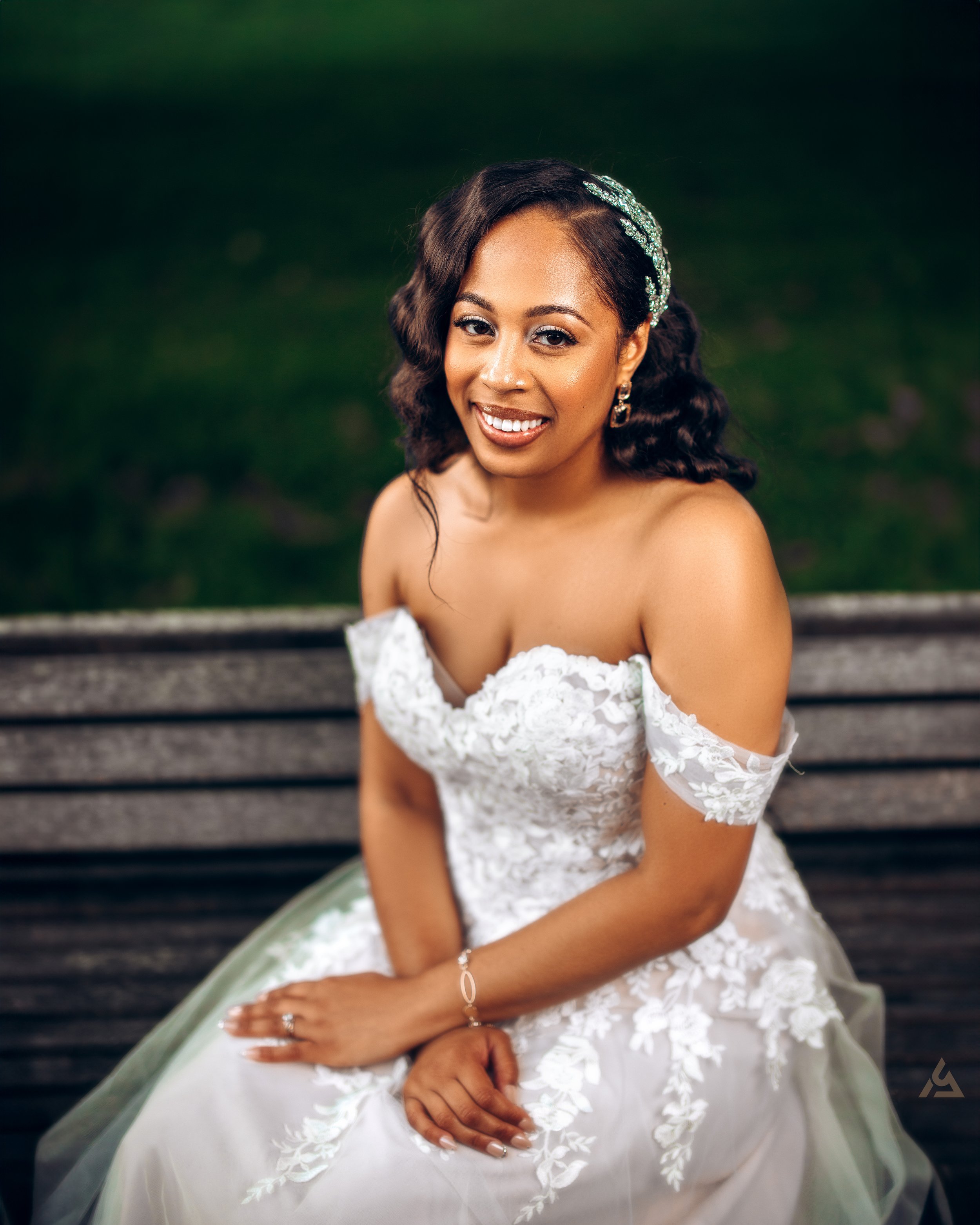 A woman wearing an off-shoulder white lace wedding dress sitting on a wooden bench outdoors, smiling at the camera.