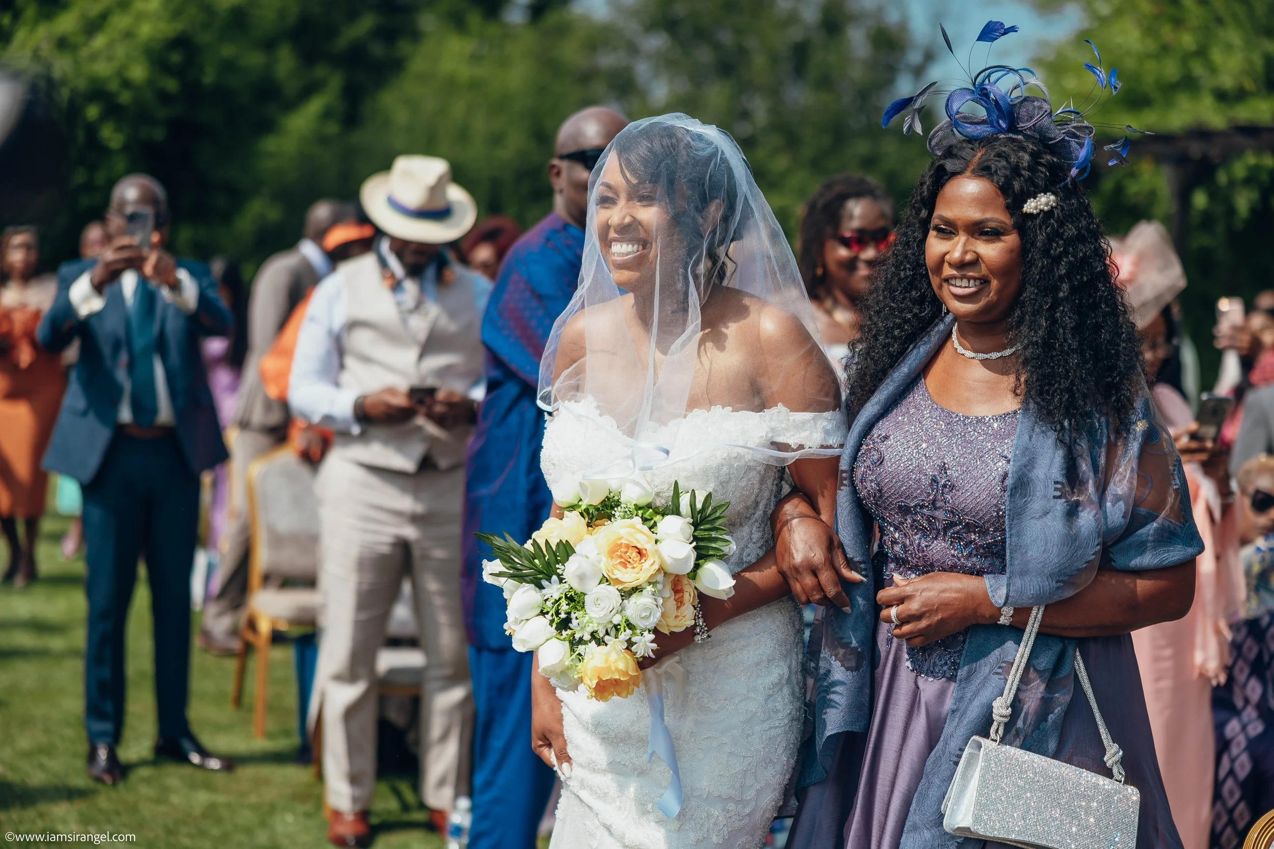 A wedding procession outdoors with the bride in a white dress, holding a bouquet, and smiling as she walks with a woman in a purple and blue dress, both wearing accessories. Guests in colorful attire are in the background.
