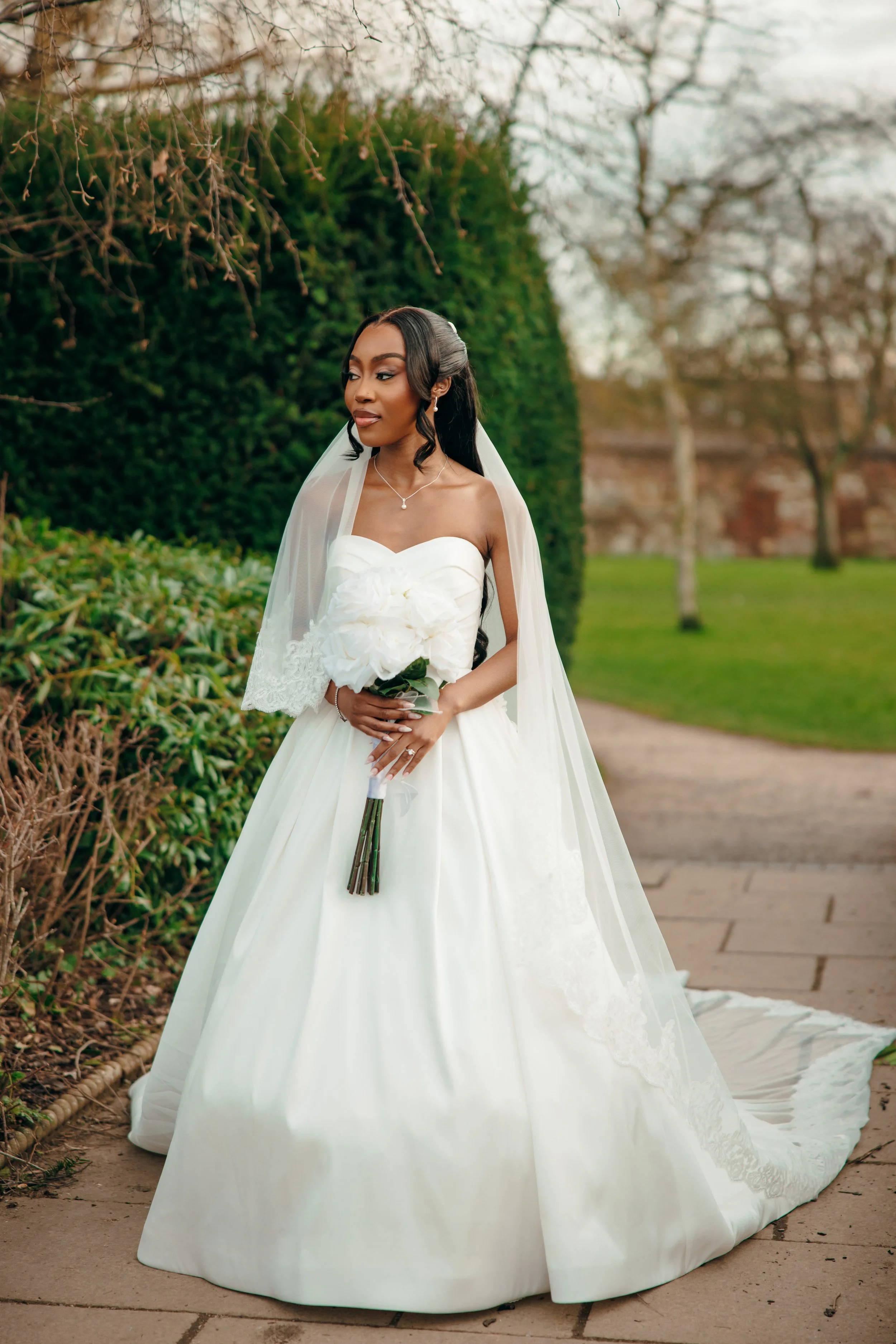 A bride in a white wedding gown holding a bouquet of white flowers outdoors, with greenery and trees in the background.