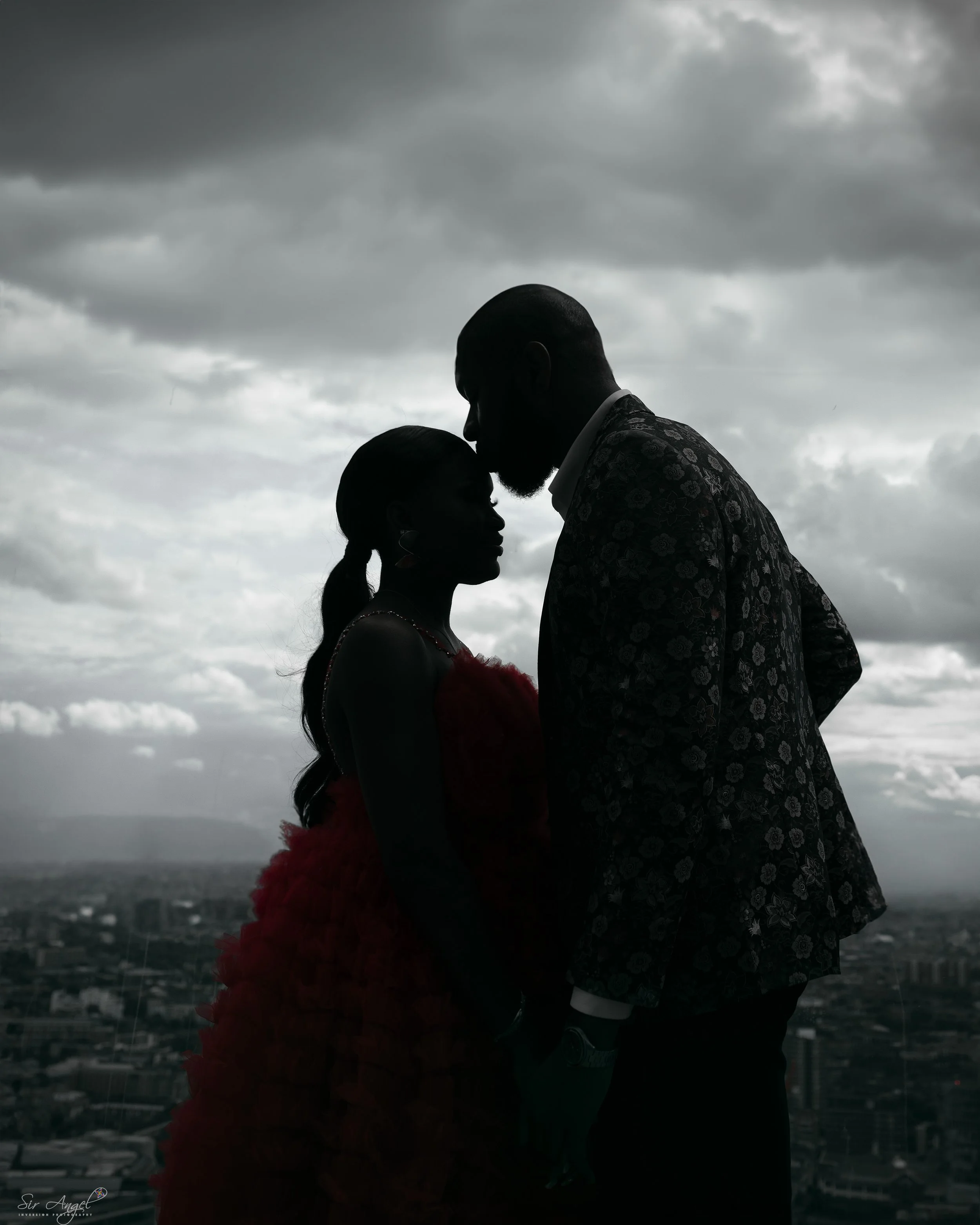 Silhouettes of a man and woman facing each other, with the man gently touching their foreheads together, standing against a cloudy sky over a cityscape.