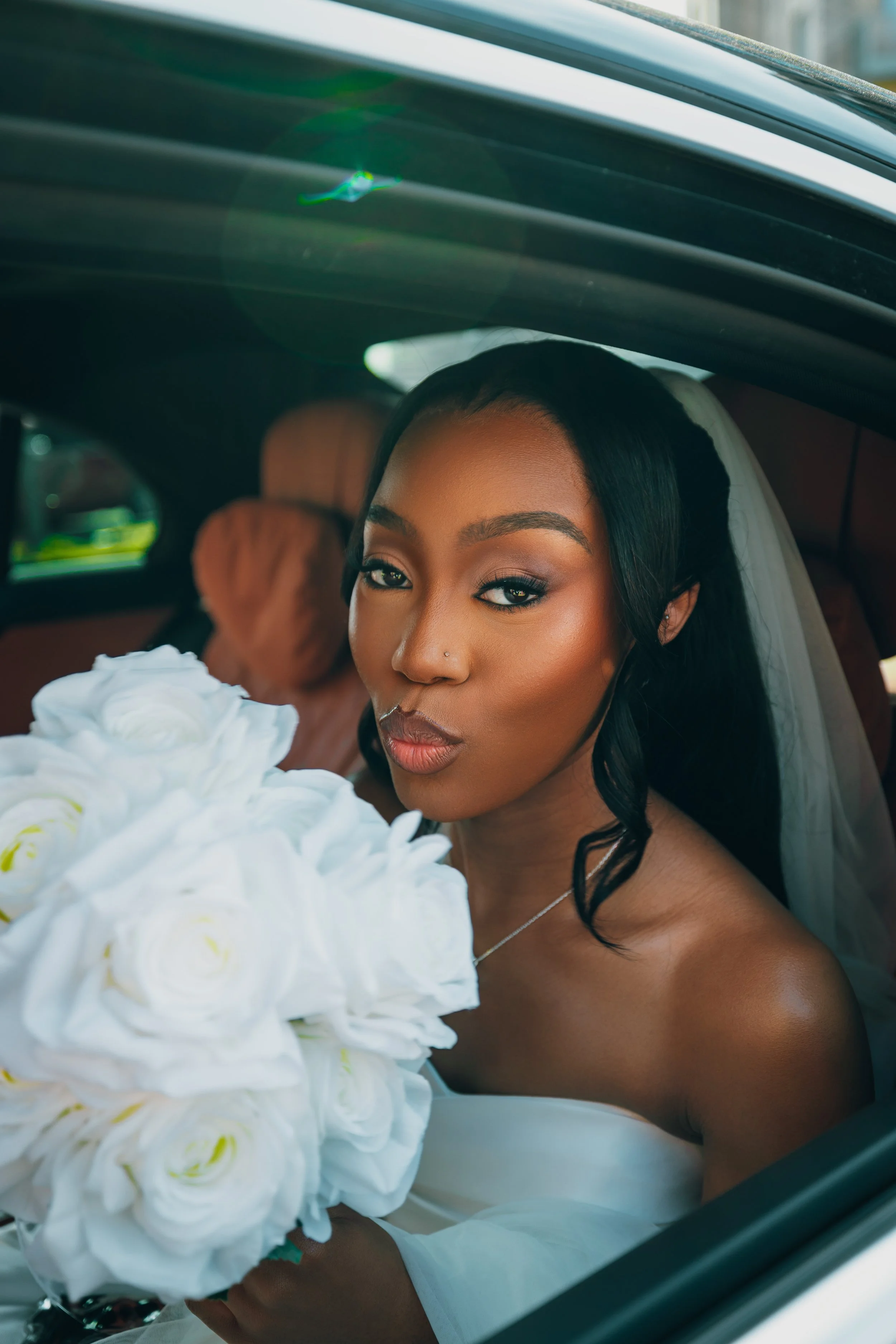 A bride sitting inside a car, holding a large bouquet of white flowers, wearing a wedding dress and veil, with makeup and jewelry, looking at the camera.