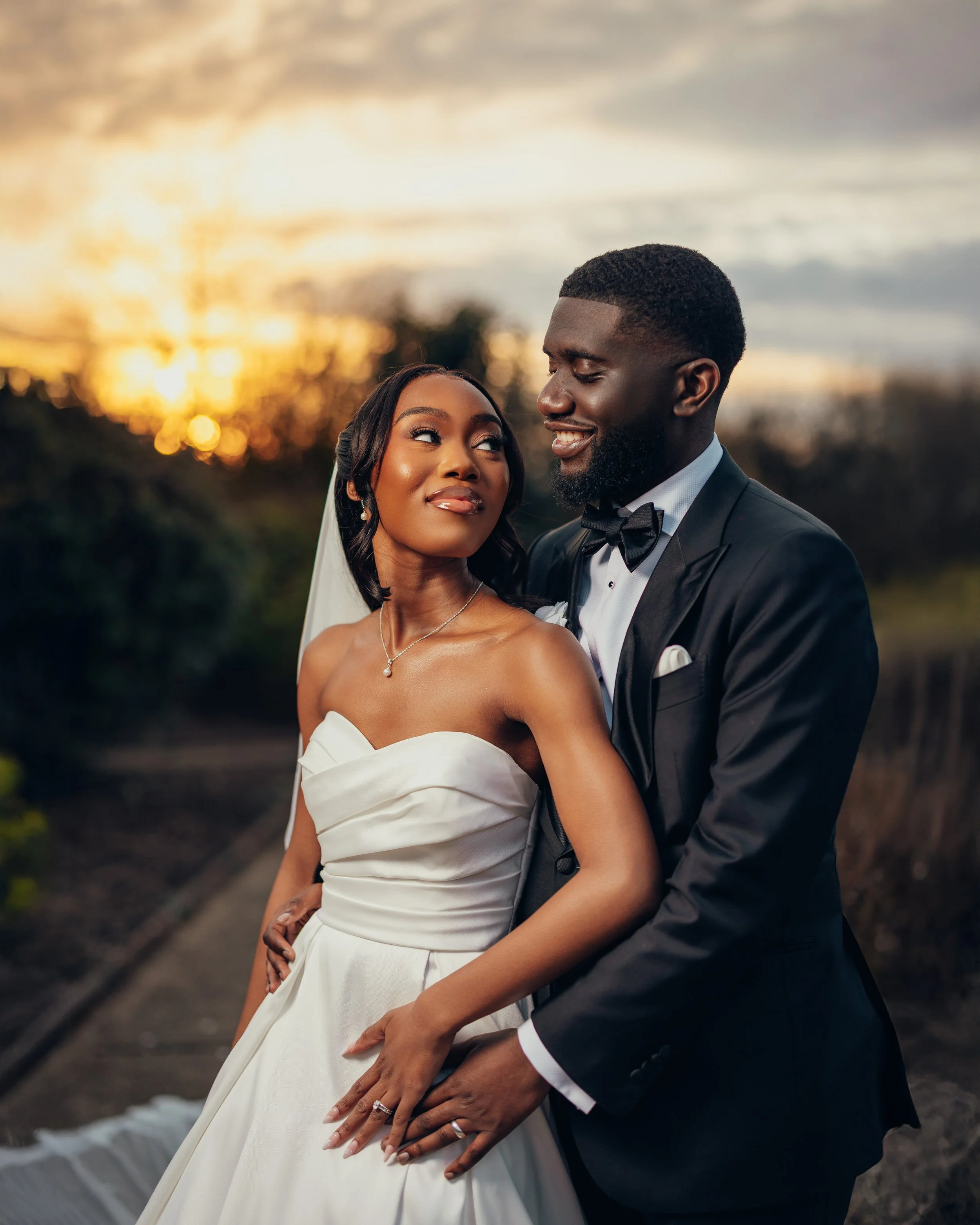 A bride and groom embrace outdoors at sunset, with the sunset sky and trees in the background. The bride wears a strapless white wedding dress and a veil, while the groom is dressed in a black tuxedo with a bow tie.